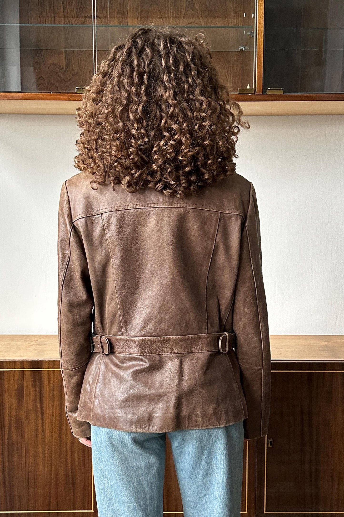 a person with curly hair wearing a brown leather jacket, standing in front of a wooden cabinet.