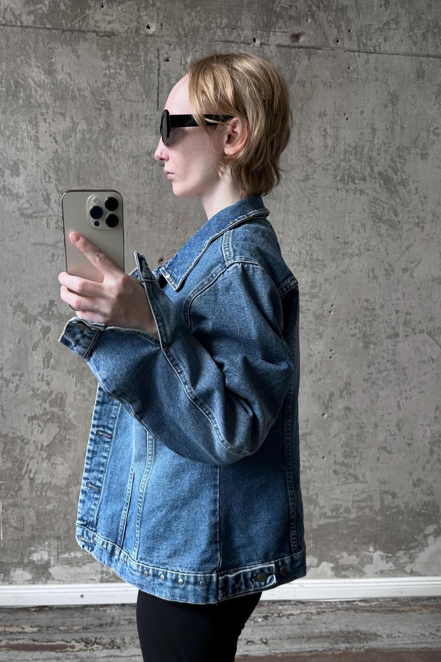 A person wearing a denim jacket and sunglasses is holding a smartphone and looking at it.