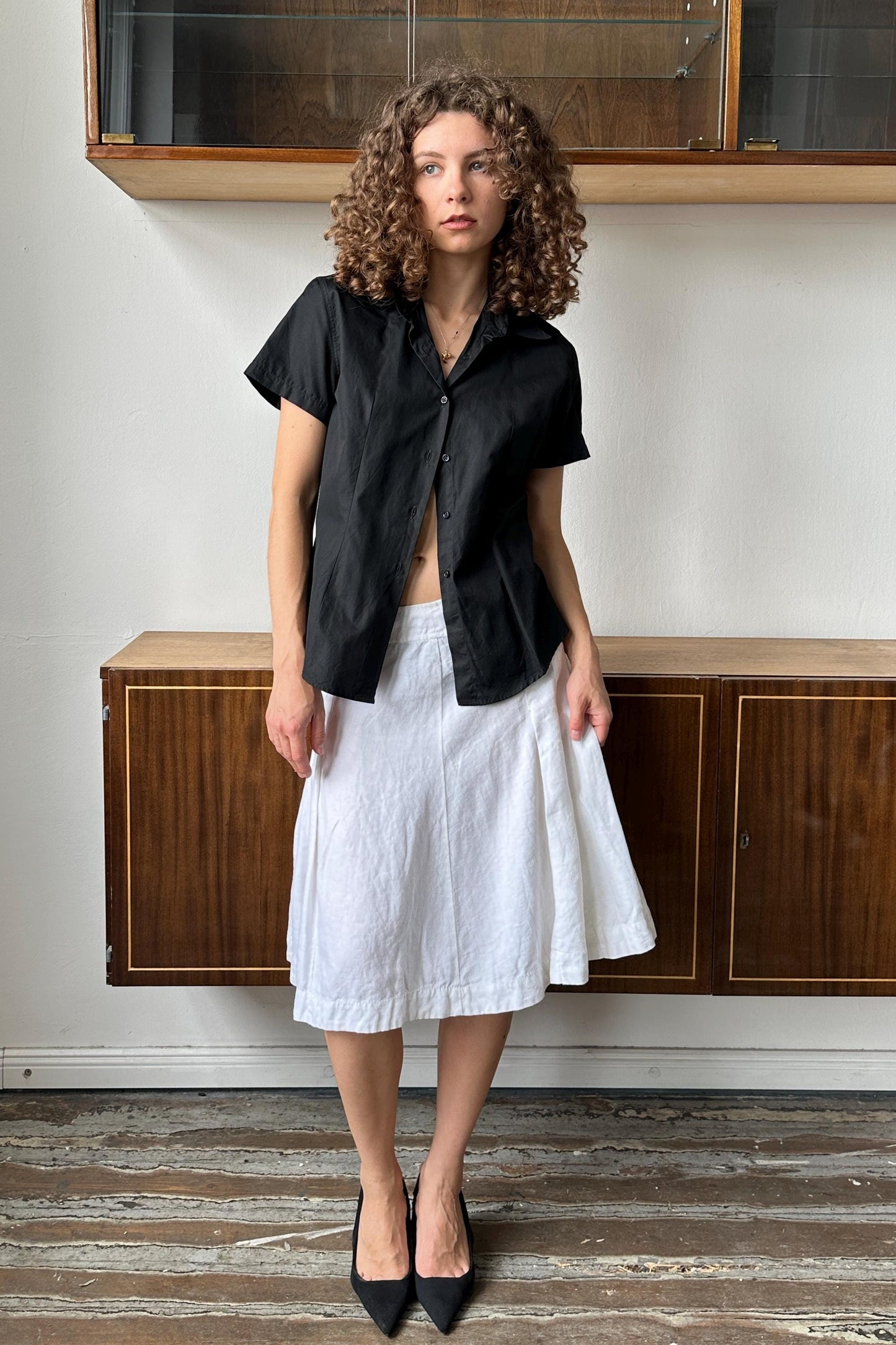 A woman with curly hair wearing a black shirt and white skirt stands in a room with wooden cabinets.