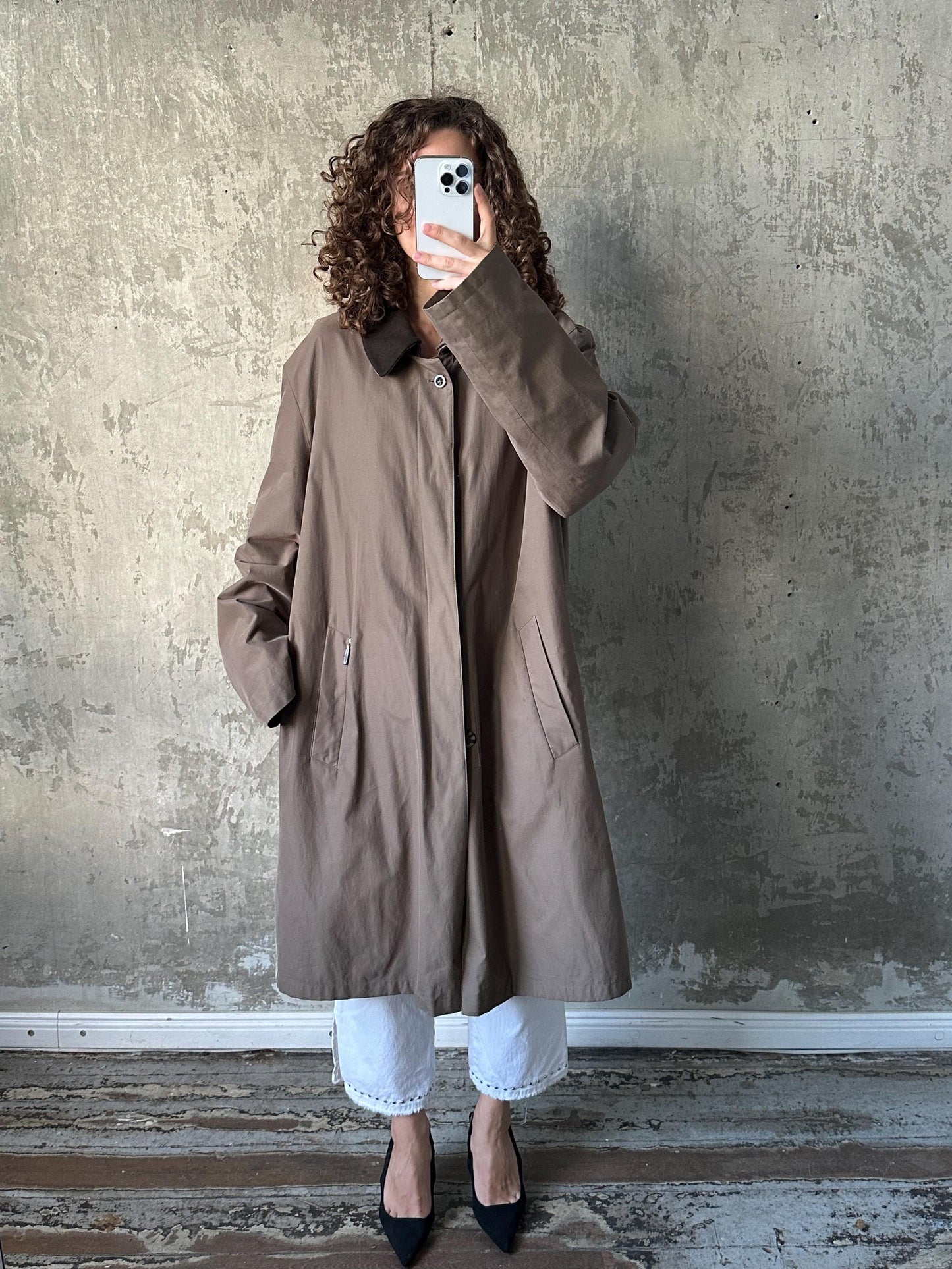 A person with curly hair is taking a selfie with a smartphone, wearing a brown coat and white pants, standing in front of a concrete wall.