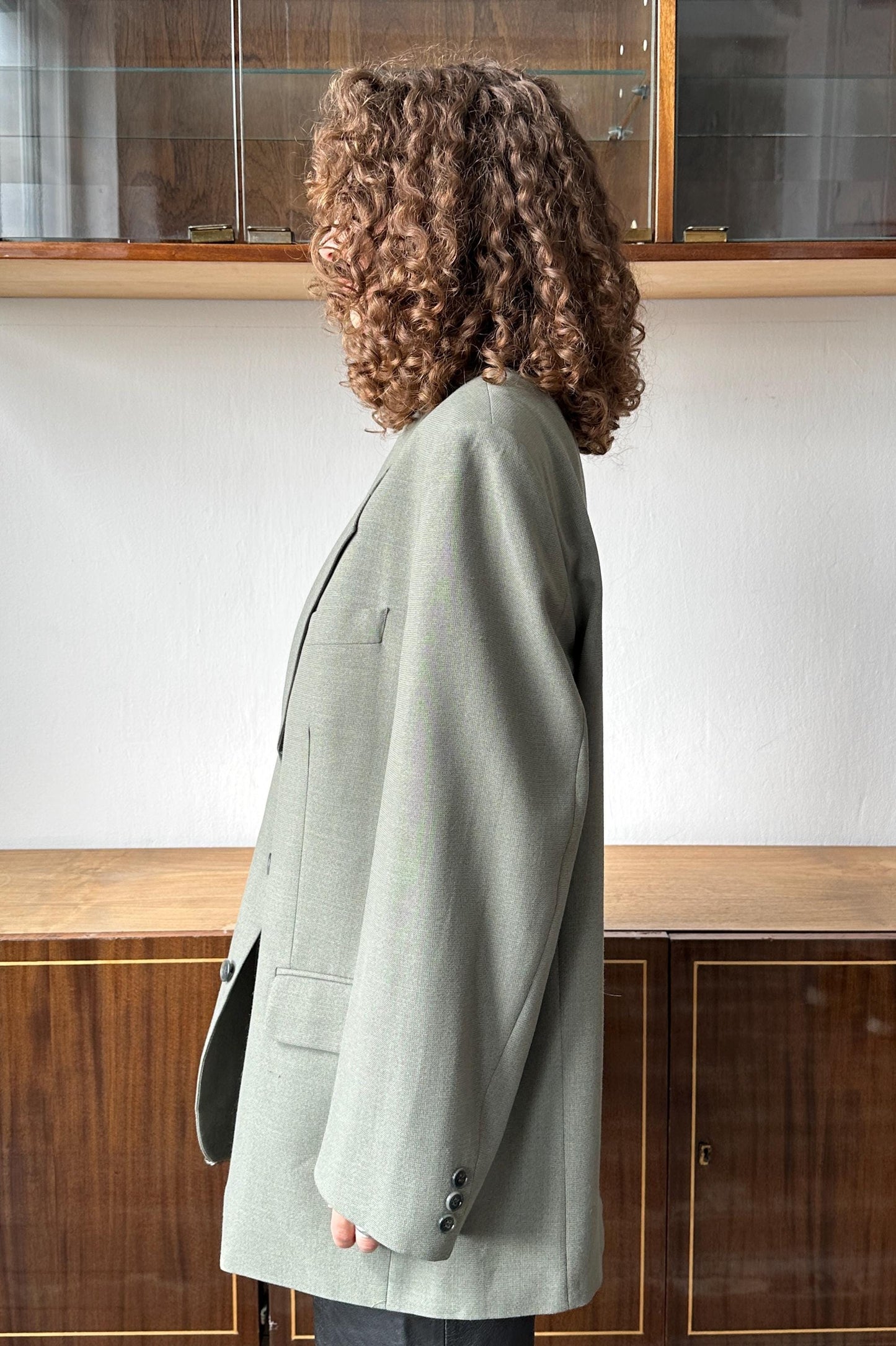 A person with curly hair wearing a light green blazer stands in front of a wooden cabinet.