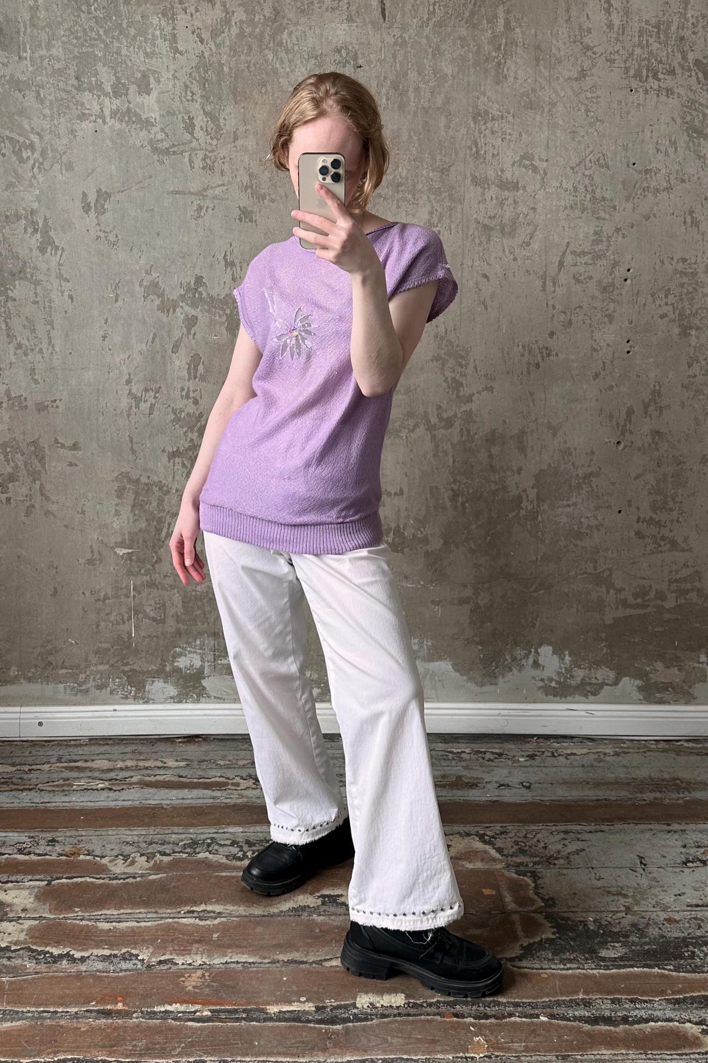 A person is taking a selfie with a smartphone, wearing a purple top and white pants, standing in front of a concrete wall.
