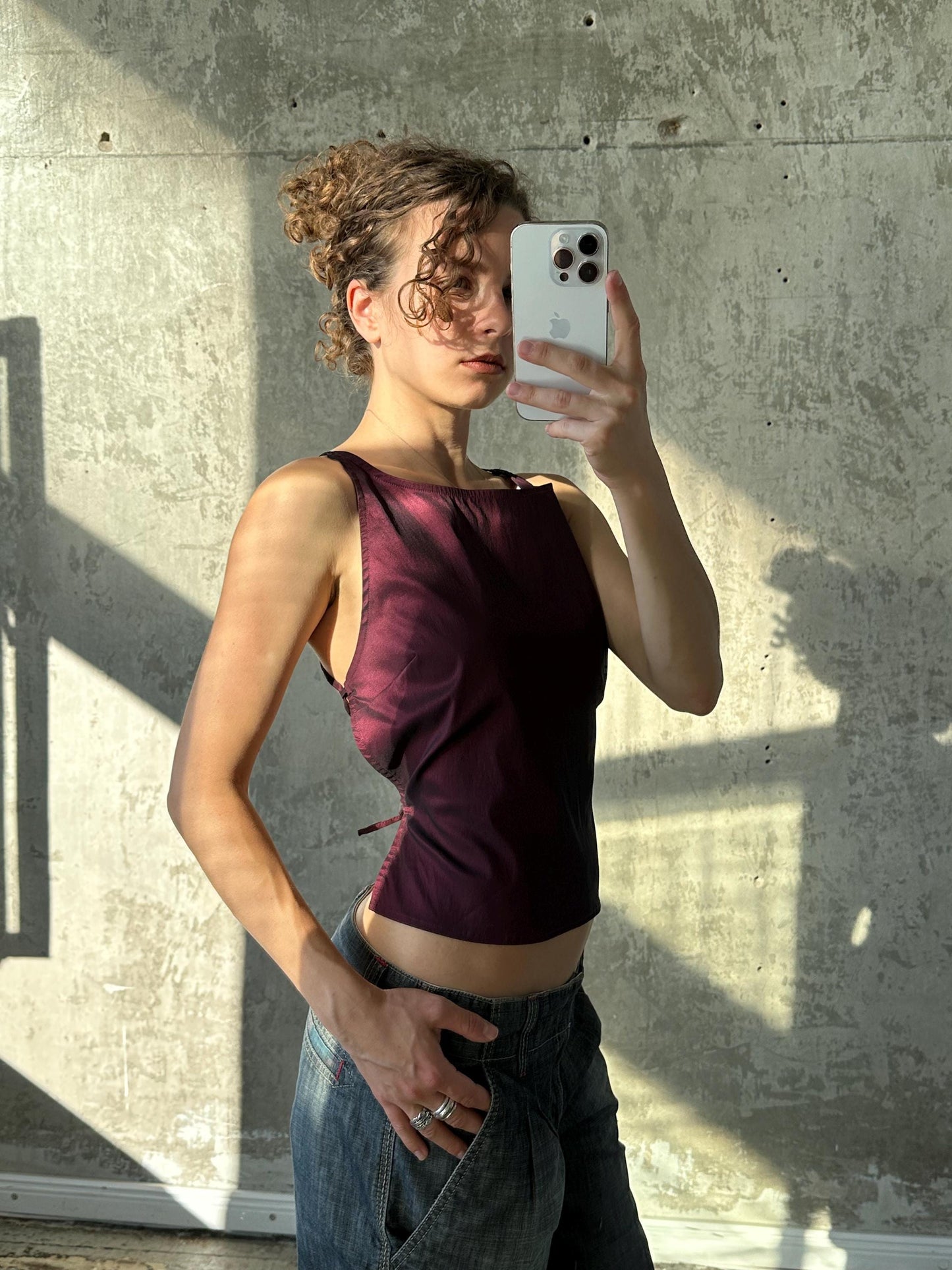 A woman in a purple top is taking a selfie with her iPhone, holding the device up to her face against a concrete wall.