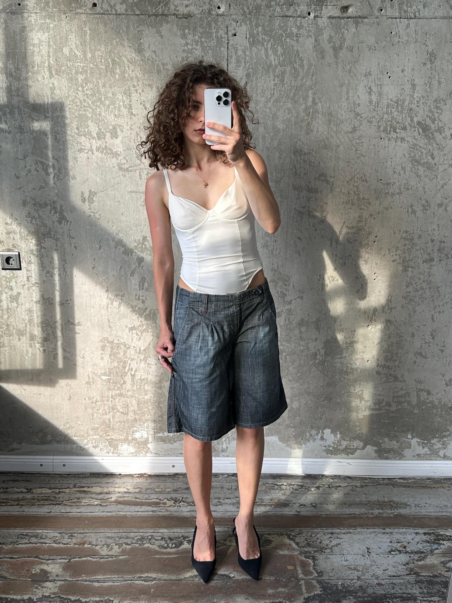 A woman with curly hair is taking a selfie with a white phone, standing in front of a concrete wall.