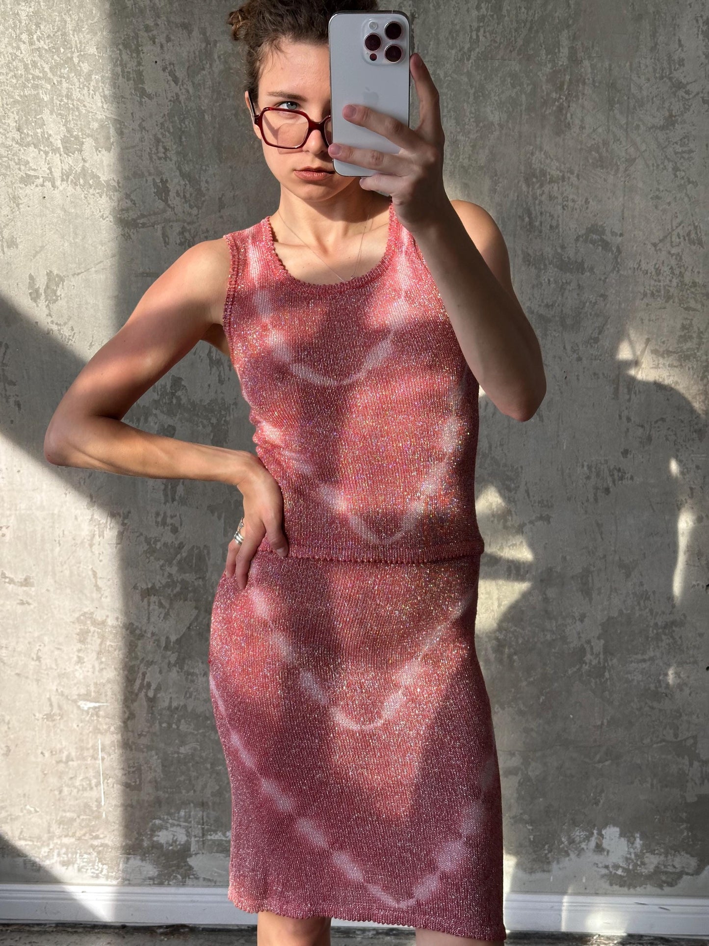 A woman wearing a pink dress is taking a selfie with a smartphone, standing in front of a concrete wall.