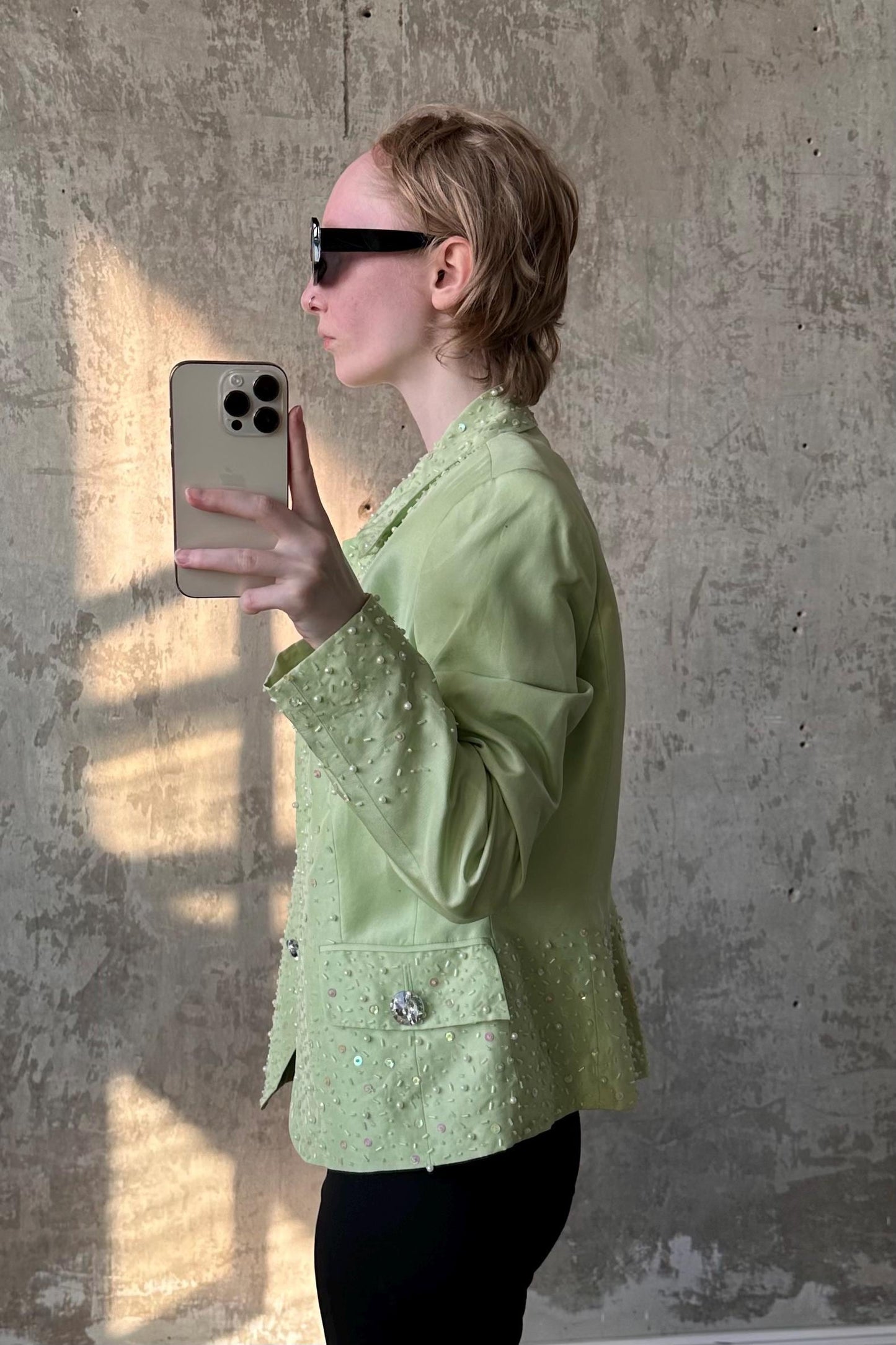 A woman in a green jacket is taking a selfie with a smartphone, holding the device up to her face and looking at the mirror.