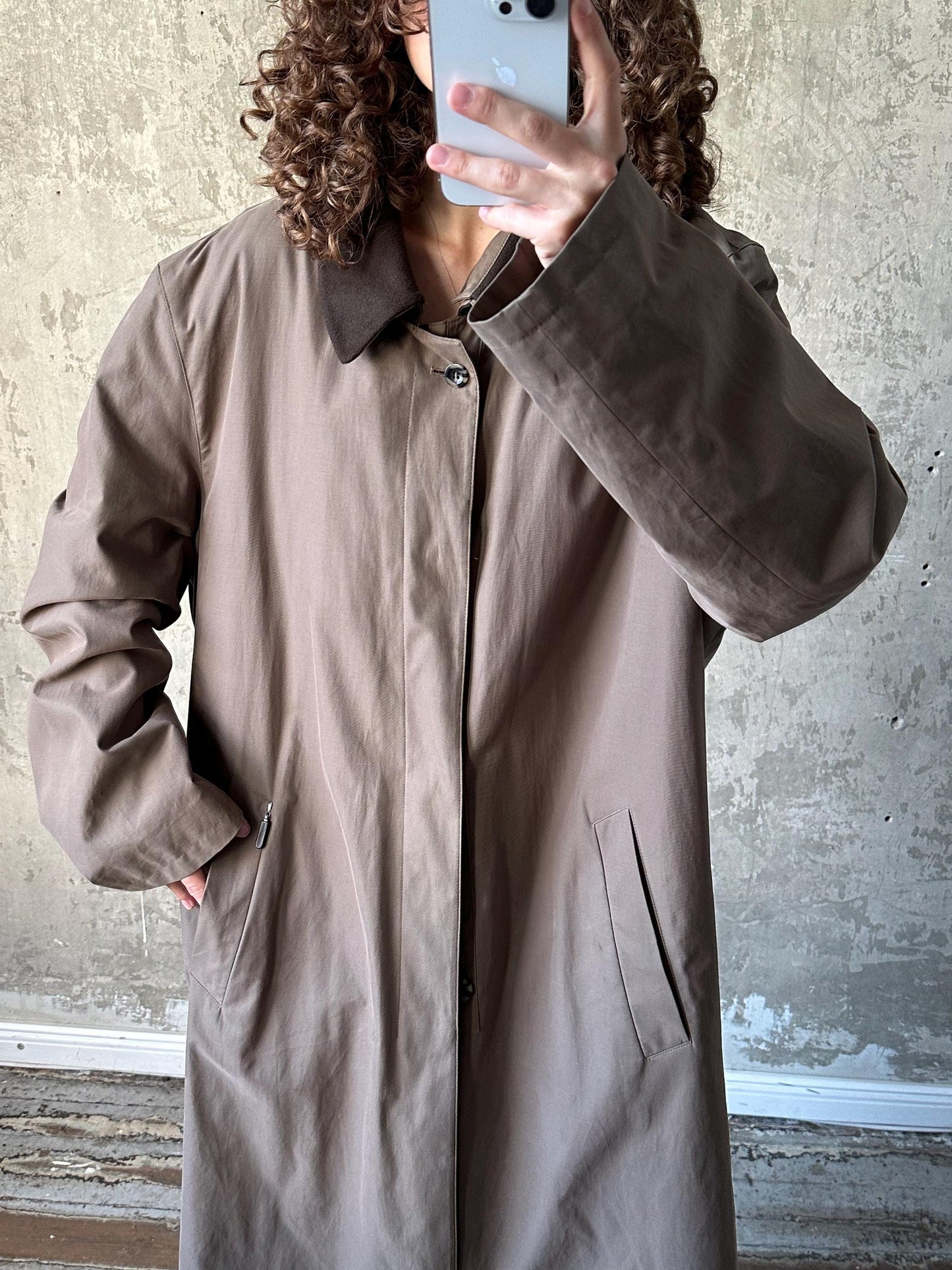 A person with curly hair is taking a selfie with a smartphone, wearing a beige coat.