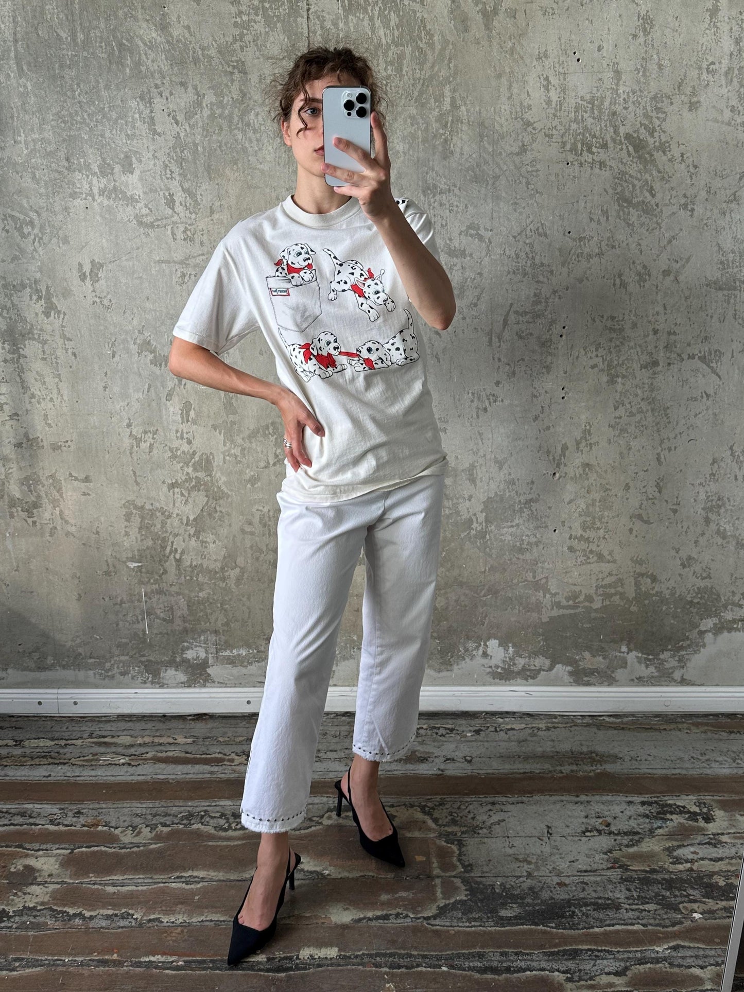 A person is taking a selfie with a smartphone, wearing a white t-shirt with a cartoon design and white pants.