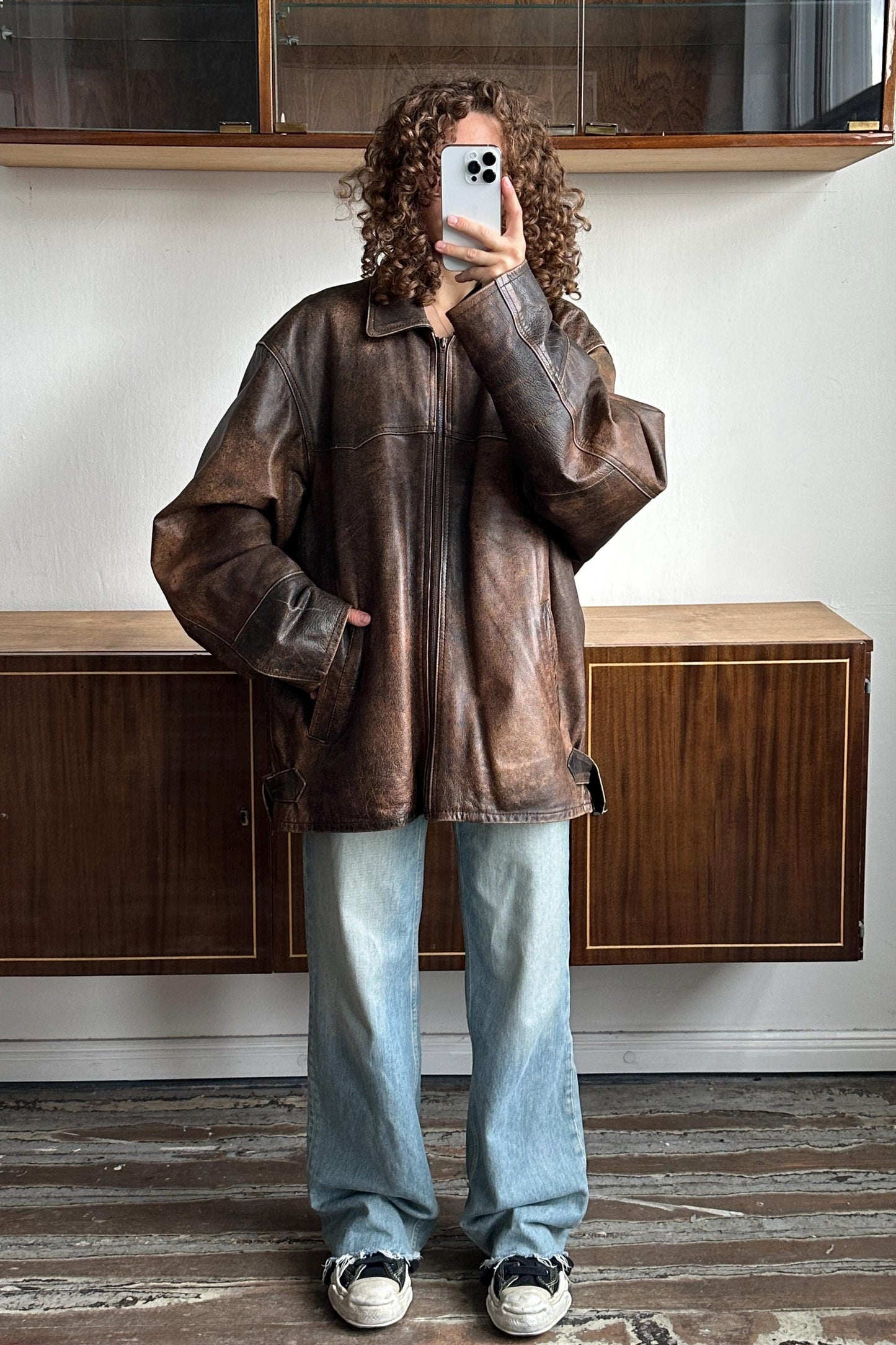 A person with curly hair is taking a selfie with a smartphone, wearing a brown leather jacket and blue jeans.