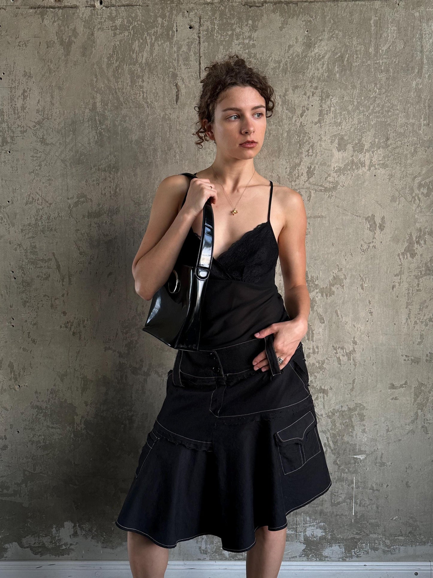 A woman in a black dress and skirt stands against a concrete wall, holding a black purse.