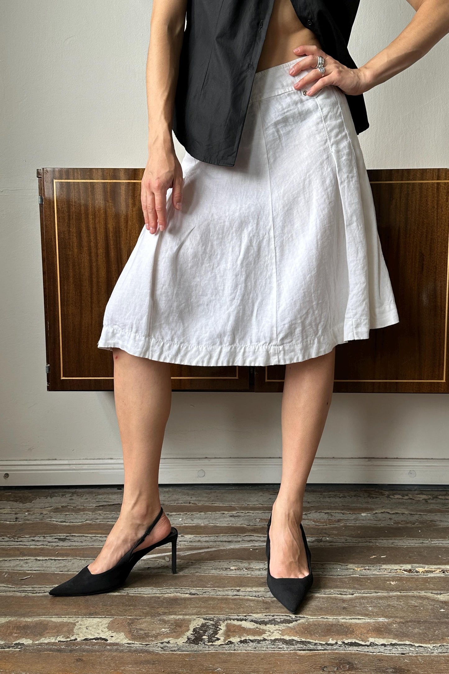 A person wearing a black vest and a white skirt stands on a wooden floor, with their hands on their hips.