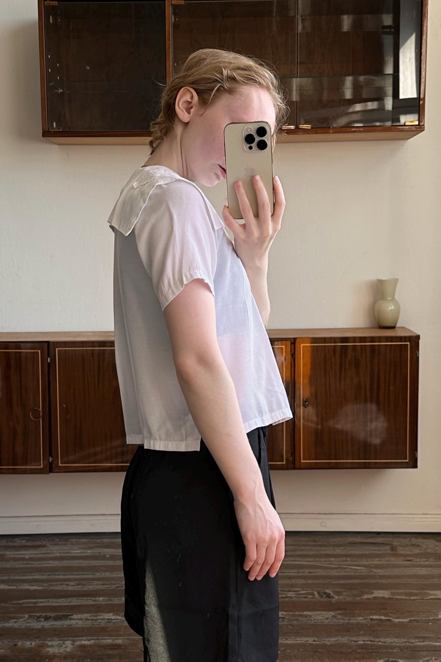 A person is taking a selfie with a white iPhone in a room with wooden cabinets and a vase on a shelf.