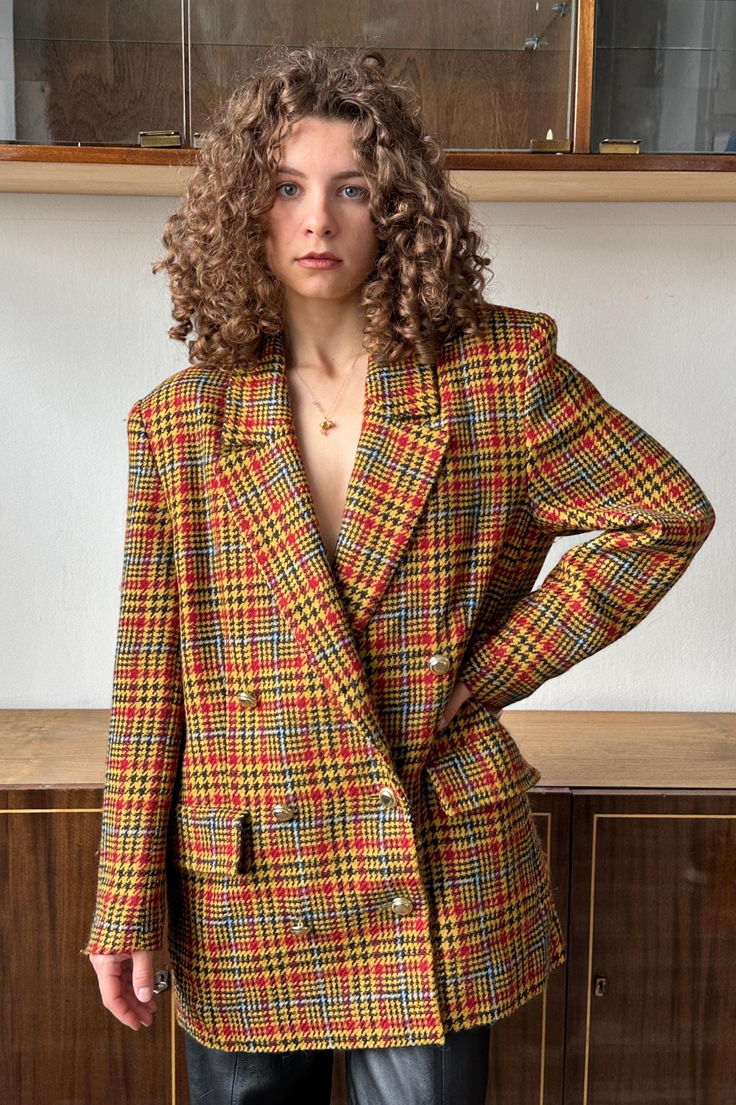 A young woman with curly hair wearing a colorful, patterned blazer stands in front of a wooden cabinet.