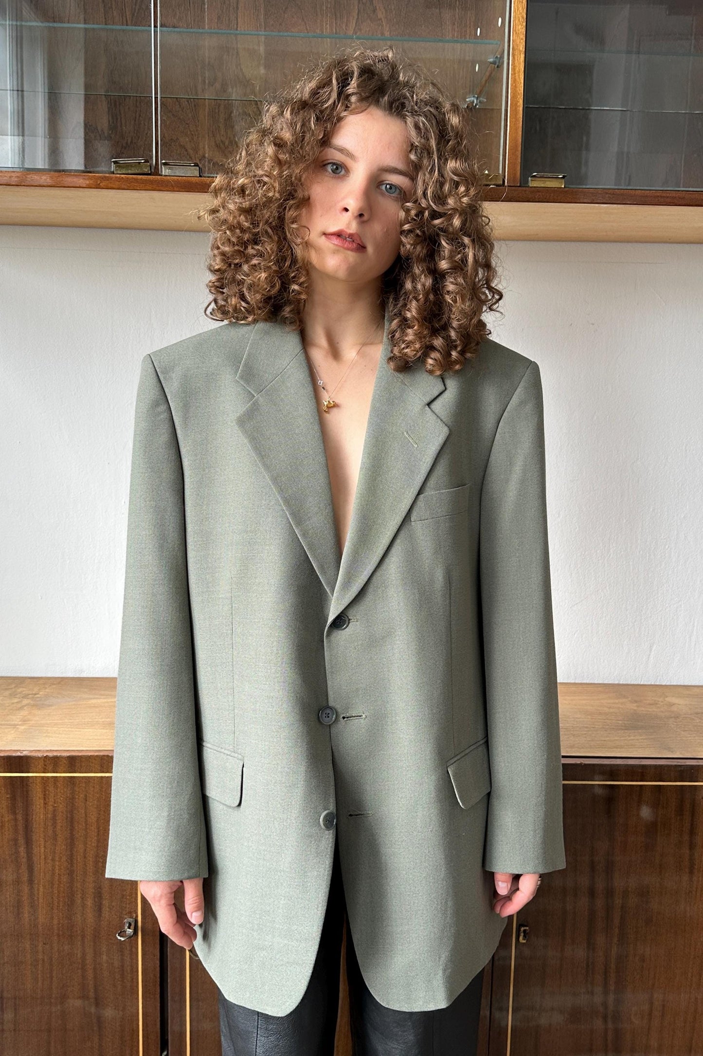 A woman with curly hair wearing a gray blazer stands in front of a wooden cabinet.