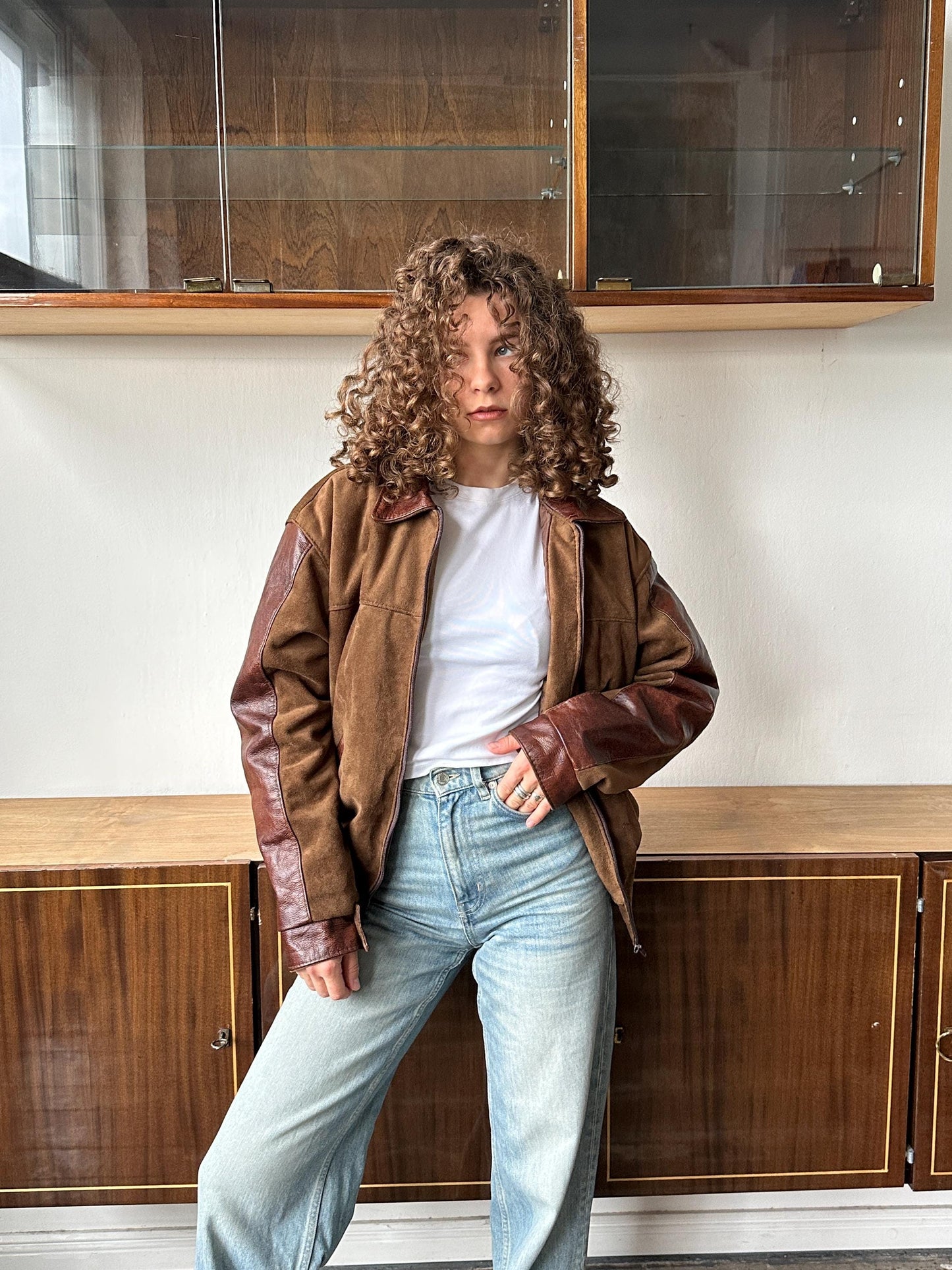 A person with curly hair wearing a brown jacket and blue jeans is standing in front of a wooden cabinet.