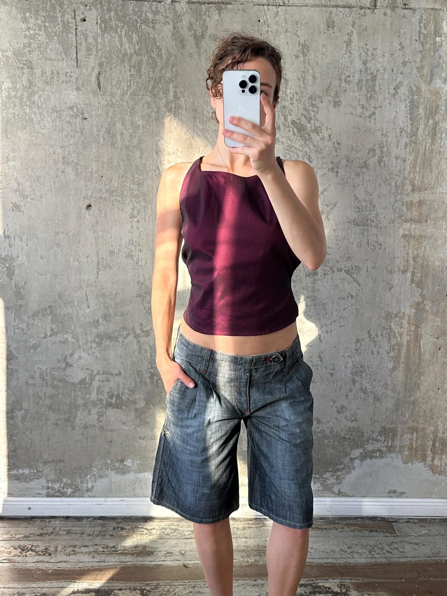 A person is taking a selfie with a smartphone, wearing a purple tank top and blue denim shorts, against a concrete wall.