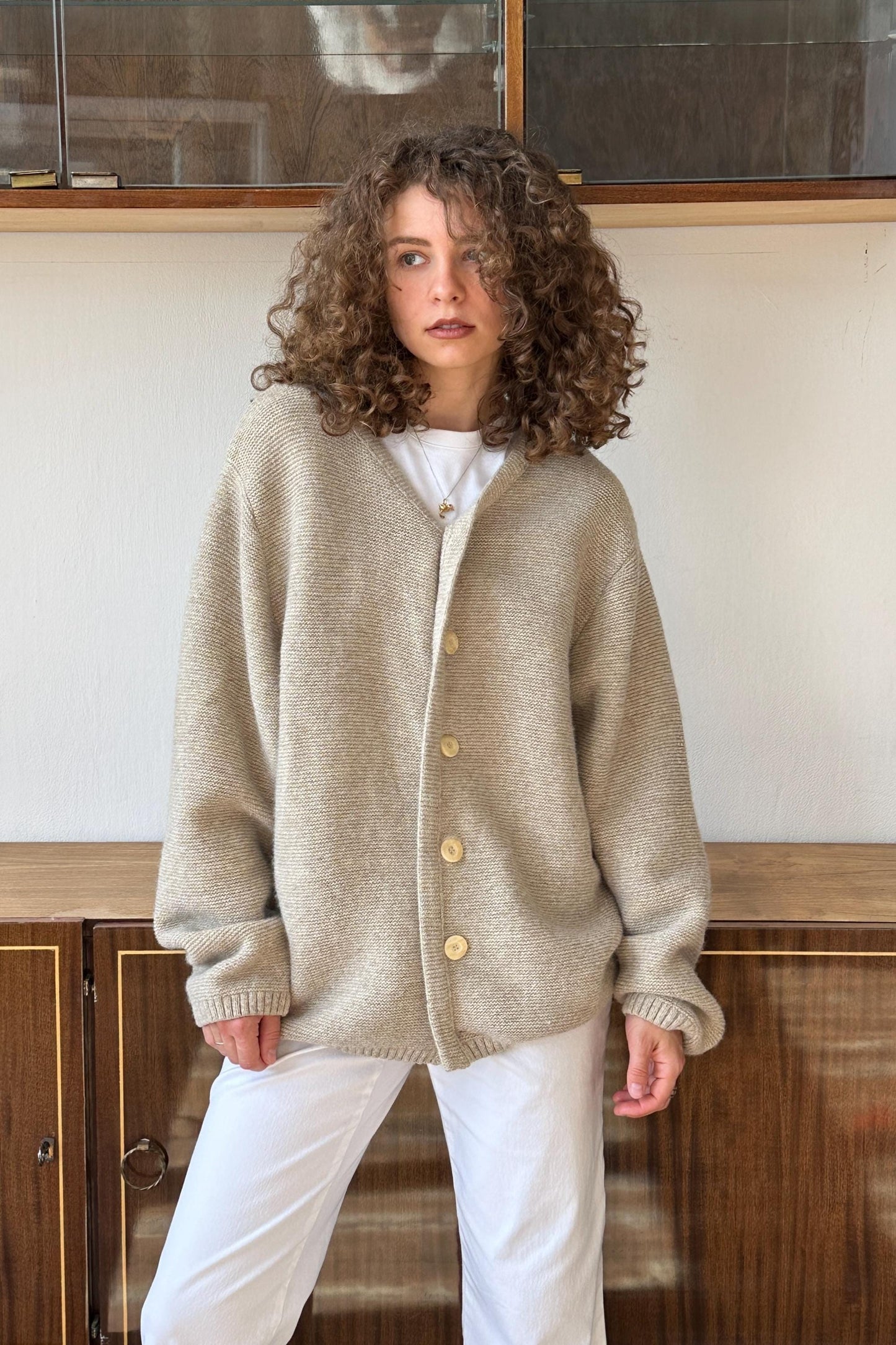 A woman with curly hair wearing a beige cardigan and white pants stands in front of a wooden cabinet.