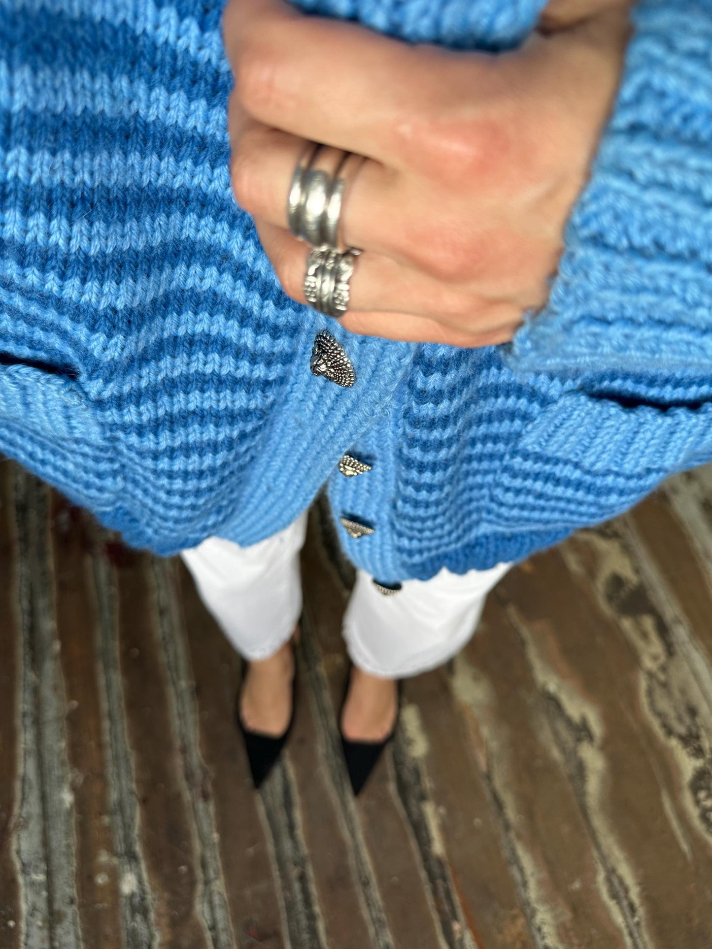 A person wearing a blue knitted cardigan and white pants, with a ring on their finger, standing on a wooden floor.
