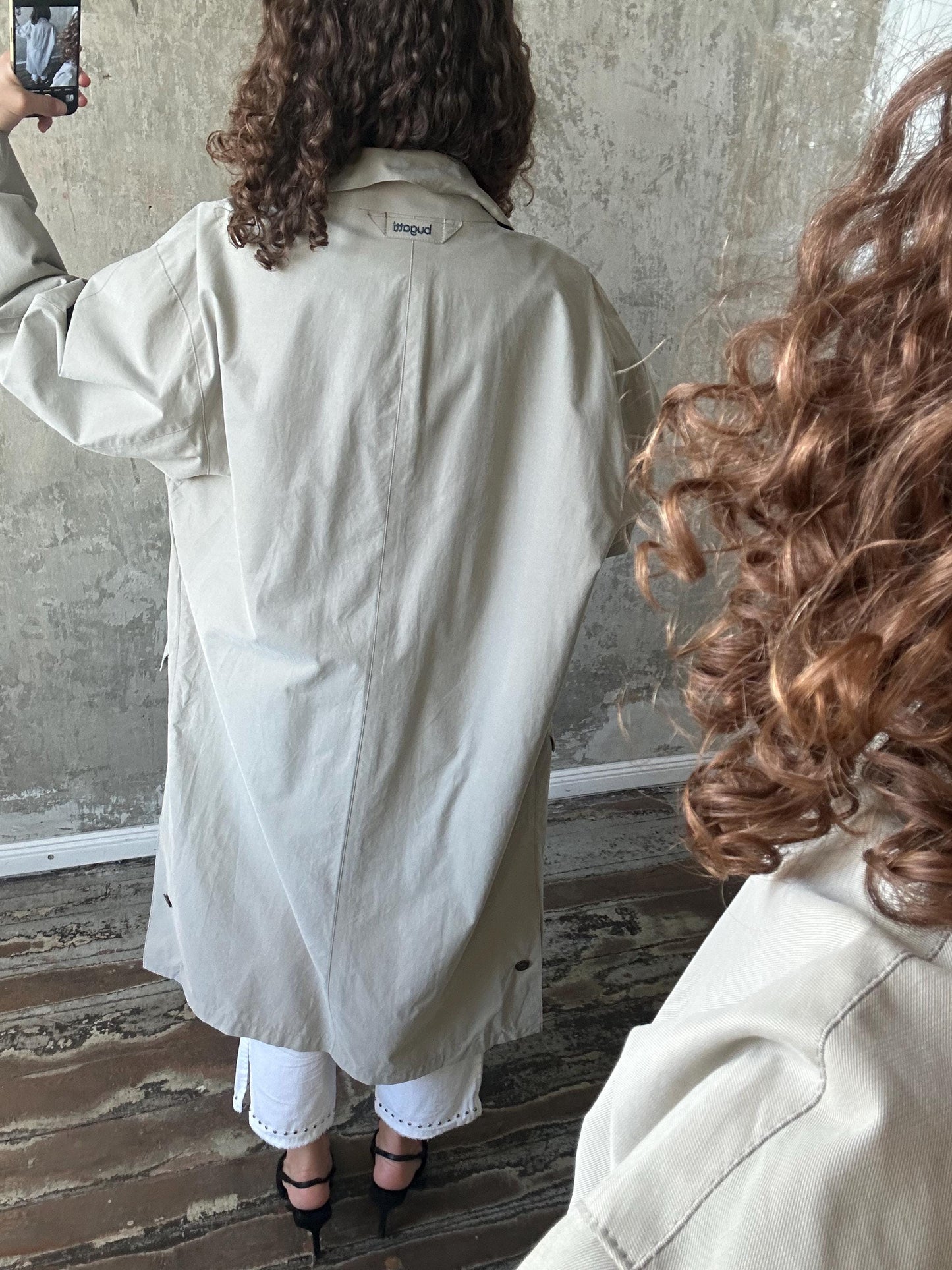 a person wearing a beige trench coat, taking a selfie with a cell phone.