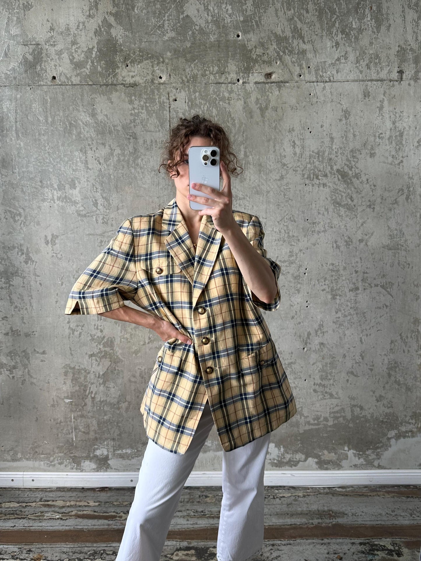 A person is taking a selfie with a smartphone, wearing a plaid jacket and white pants, against a concrete wall background.