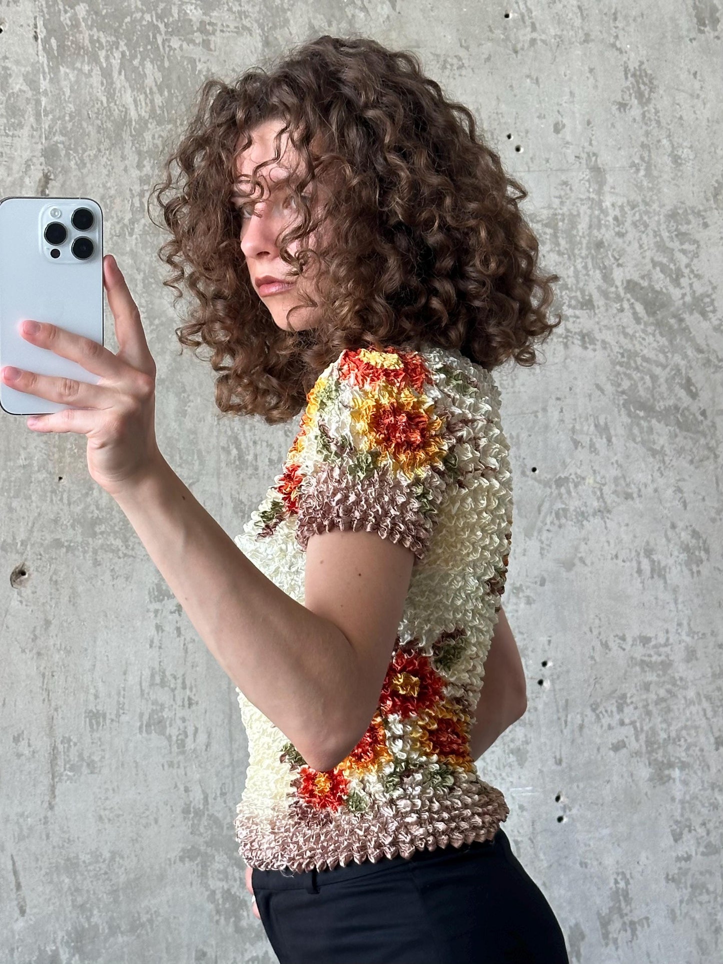 A woman with curly hair is taking a selfie with a white iPhone, wearing a colorful floral top.