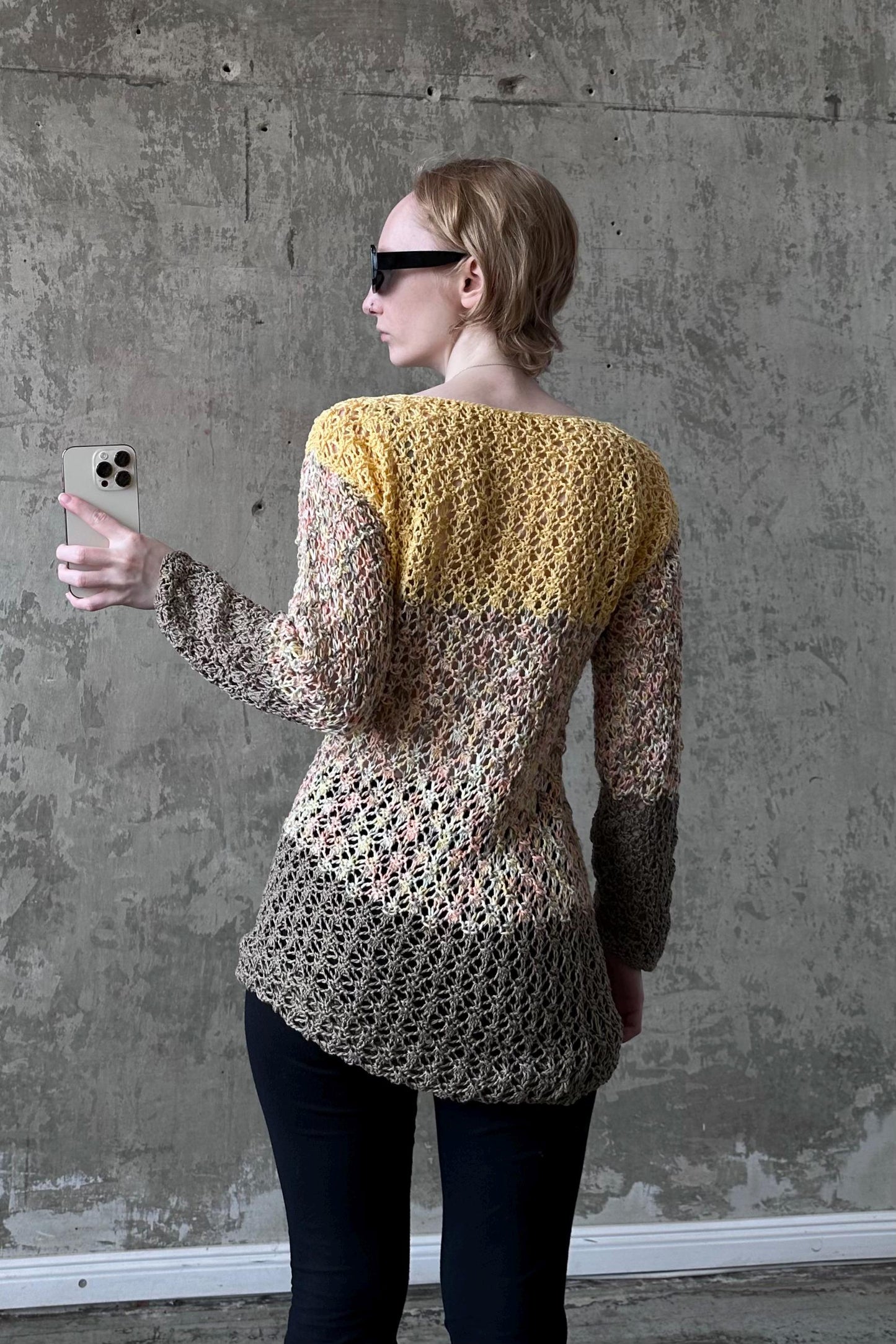 A woman wearing a yellow and brown crocheted sweater is taking a selfie with her iPhone against a concrete wall.