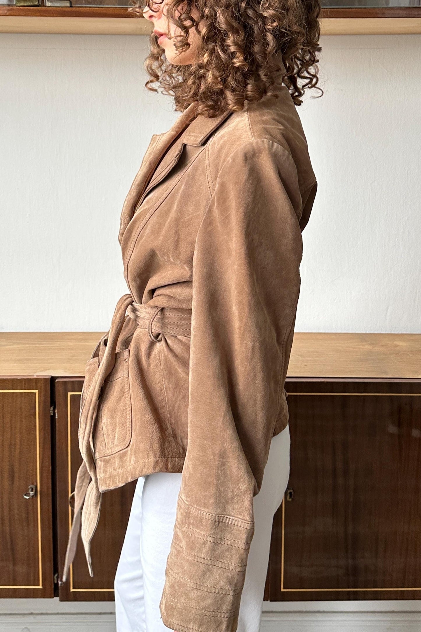 A woman with curly hair wearing a tan leather jacket stands in front of a wooden cabinet.
