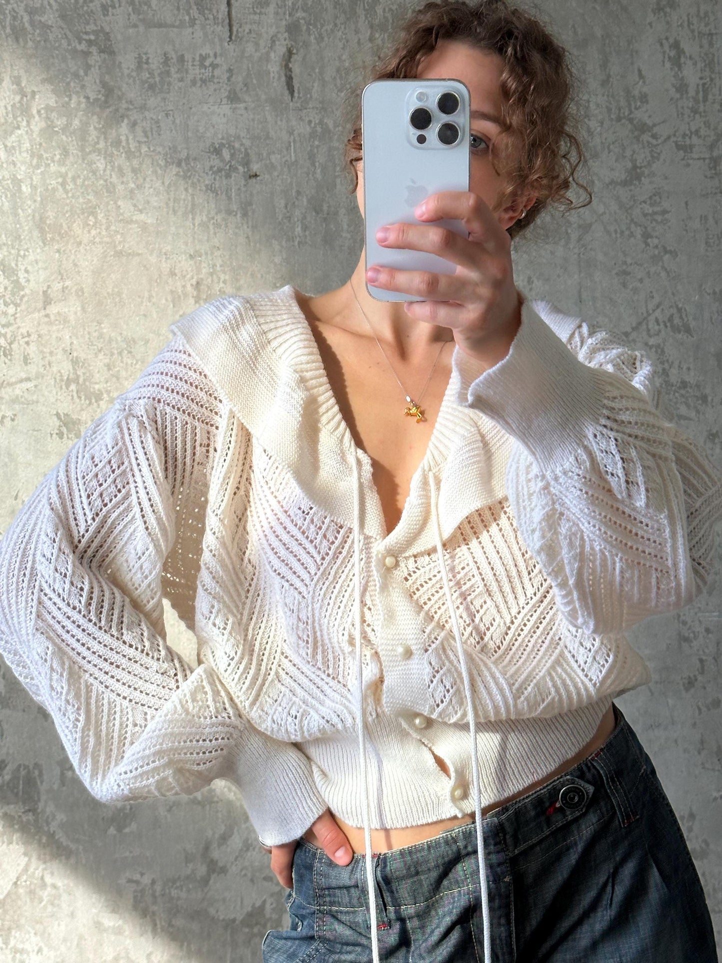 A woman is taking a selfie with her iPhone, wearing a white cardigan and jeans.