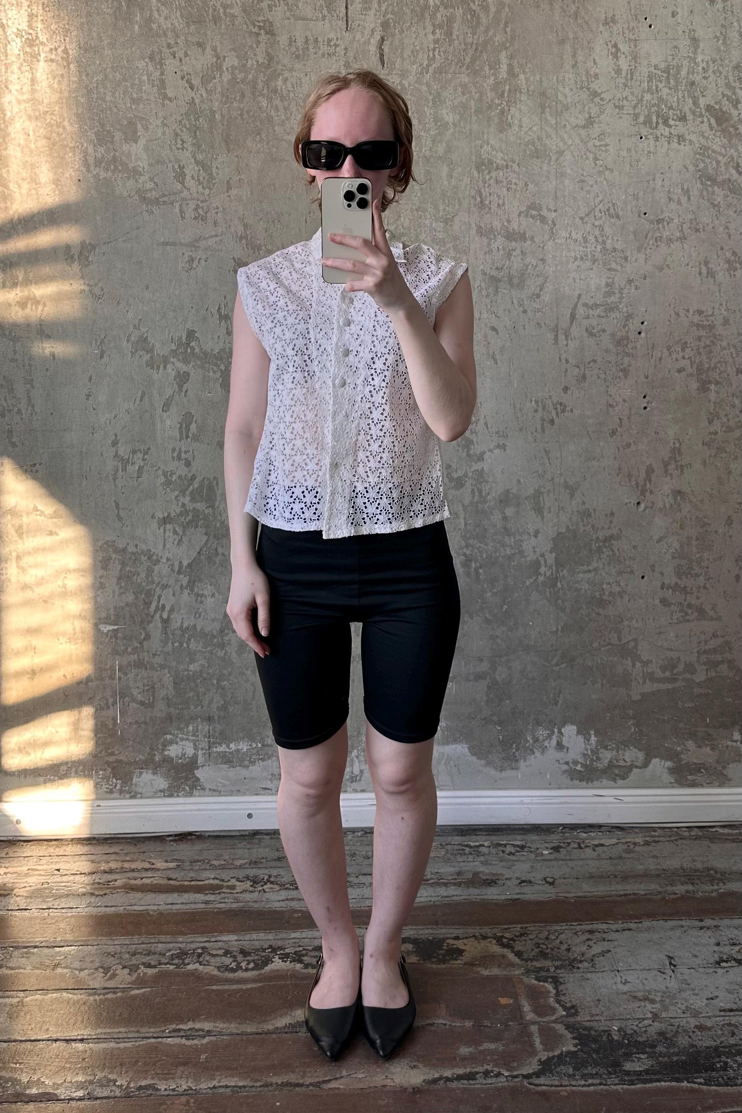 A person is taking a selfie with a smartphone, wearing a white lace top, black shorts, and black flat shoes, standing in front of a concrete wall.