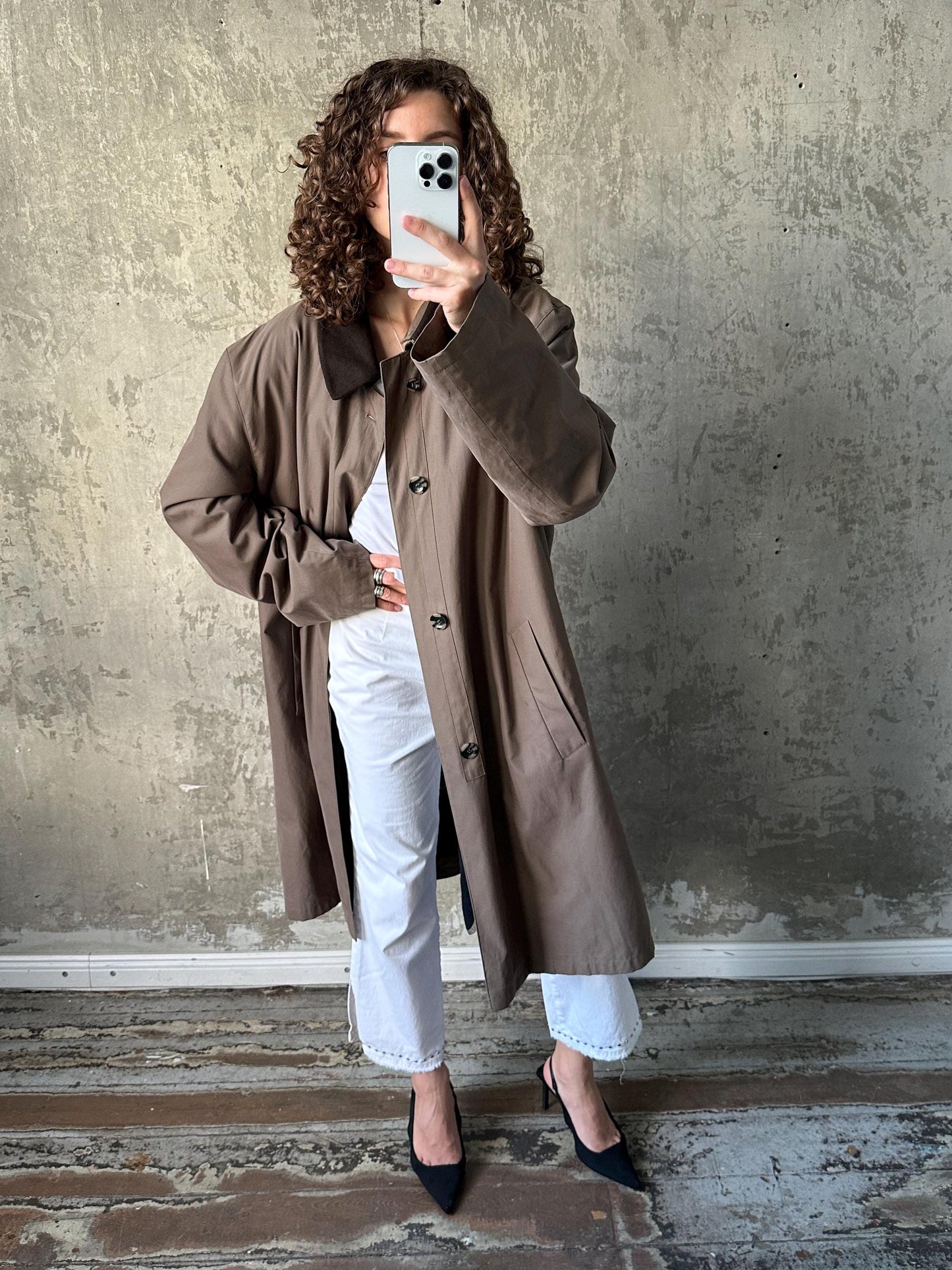 A woman with curly hair is taking a selfie with her phone, wearing a brown coat and white pants, standing in front of a concrete wall.
