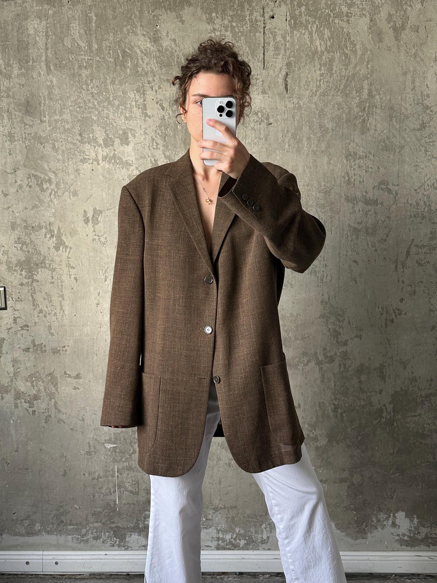 A person is taking a selfie with a smartphone, wearing a brown blazer and white pants against a concrete wall background.