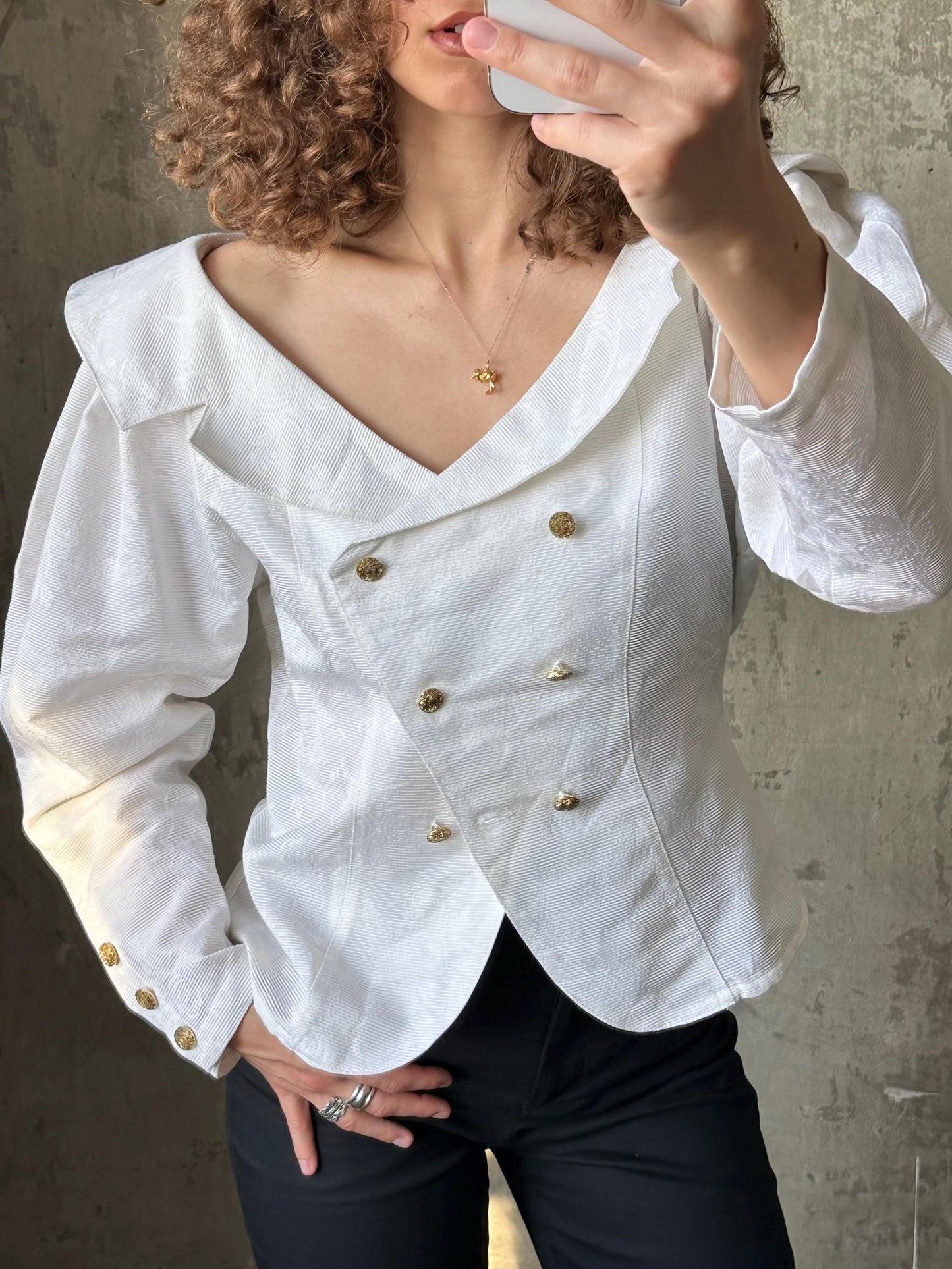 A woman with curly hair is taking a selfie with a white blouse and black pants.