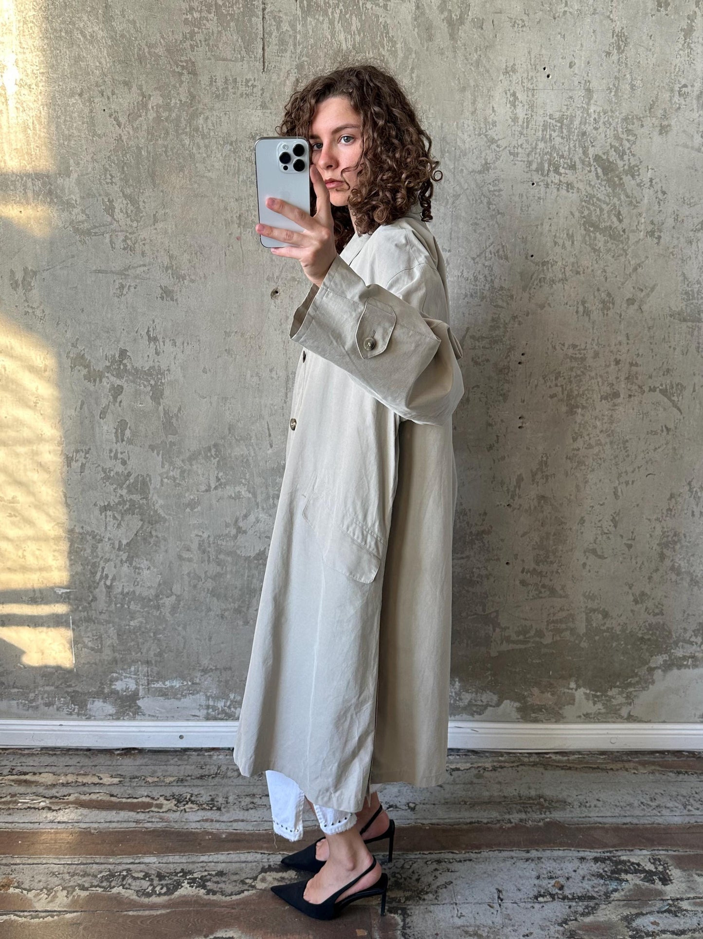A woman in a beige trench coat is taking a selfie with a smartphone, standing in front of a concrete wall.