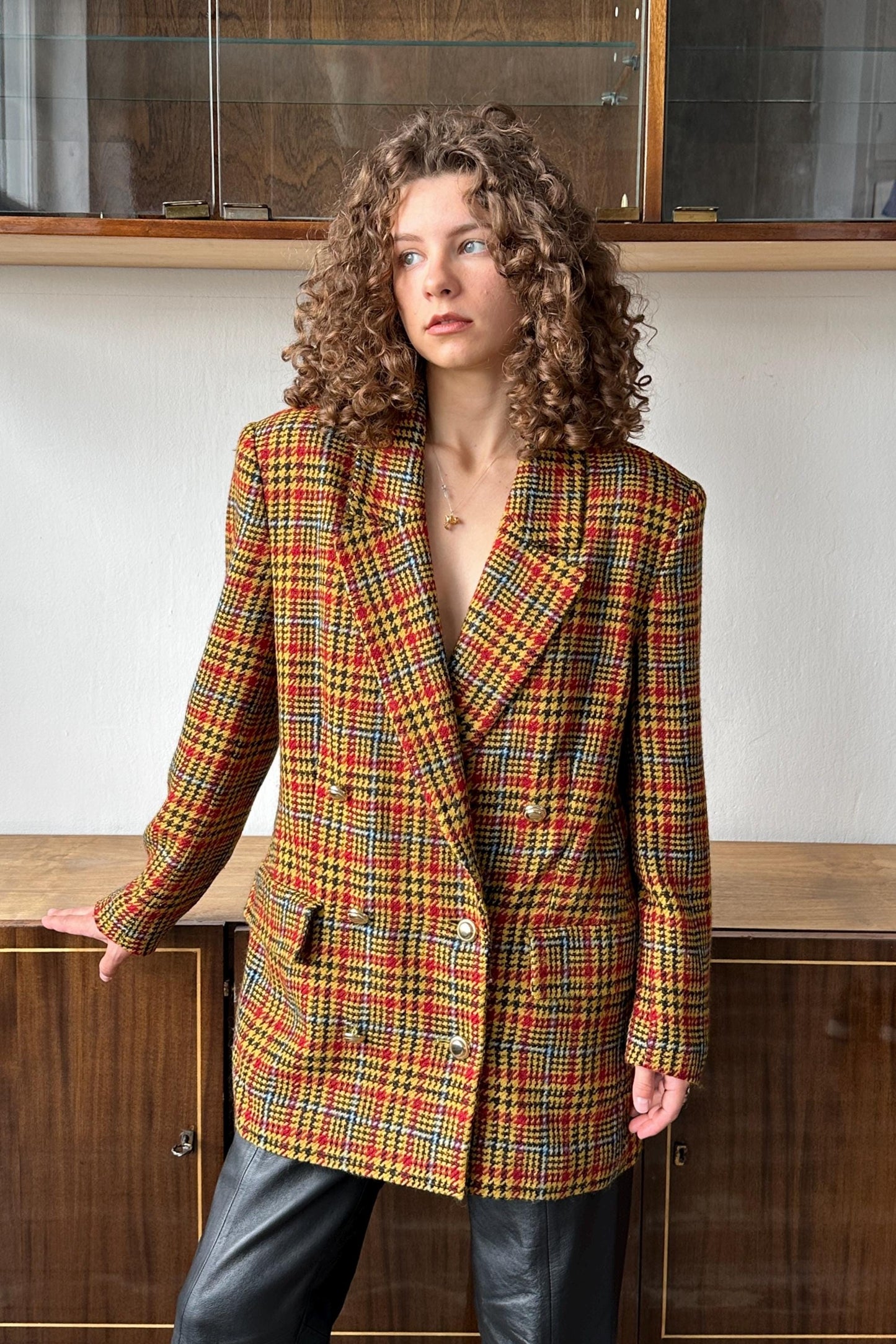 A woman with curly hair wearing a colorful, patterned blazer stands in front of a wooden cabinet.