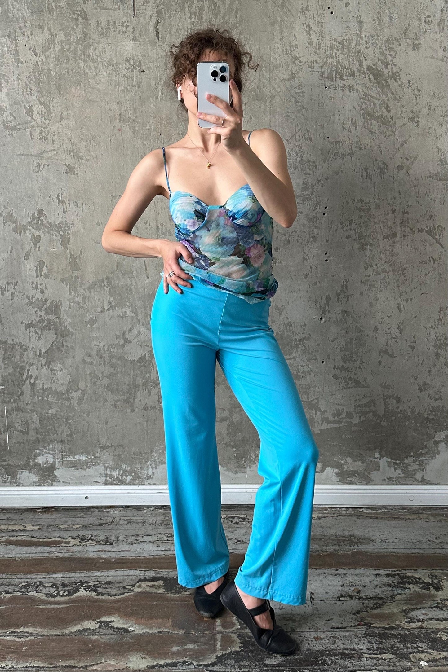 A woman is taking a selfie with her phone, wearing a blue floral top and blue wide-leg pants, standing in front of a concrete wall.