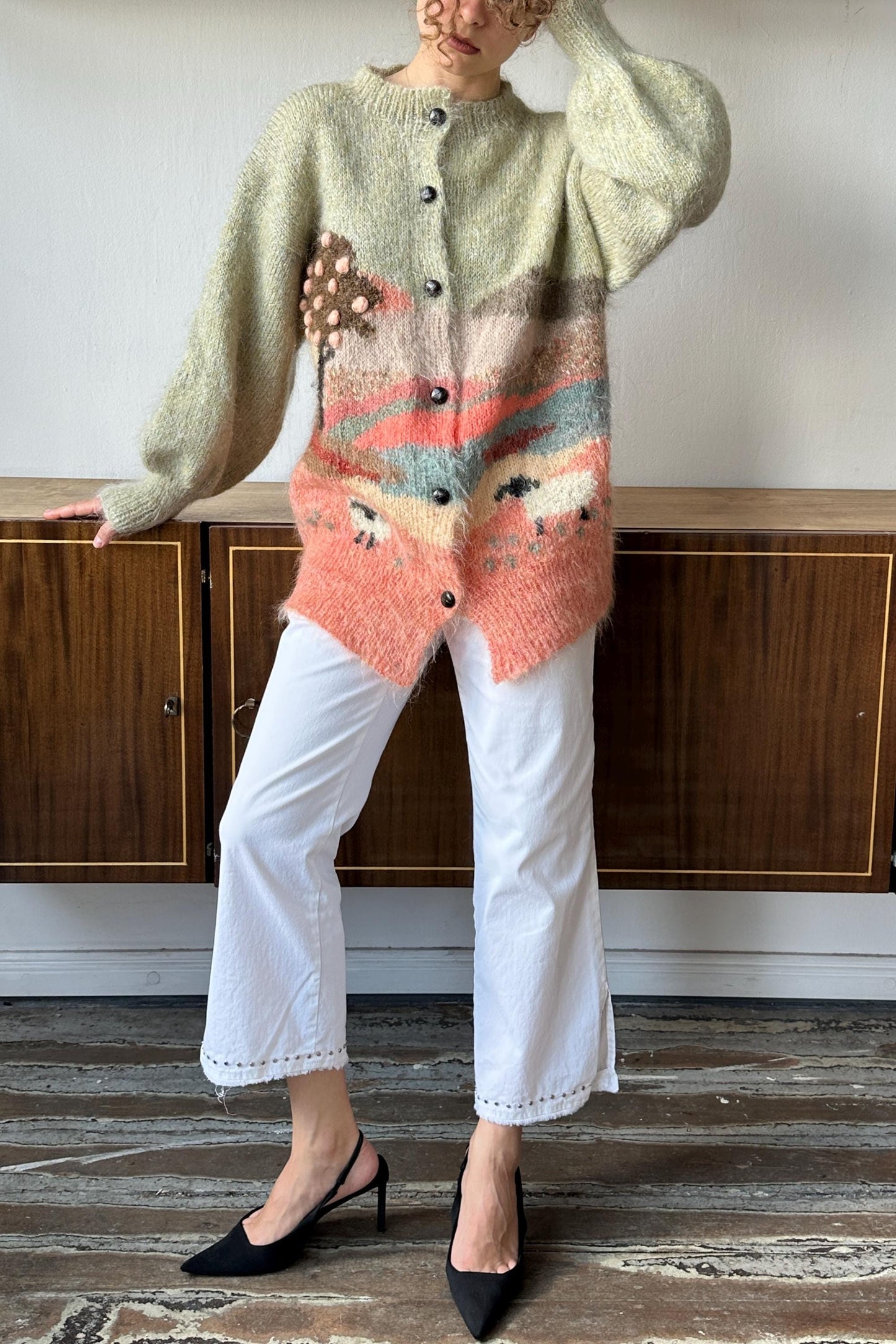 A person wearing a pink and white knitted cardigan with a colorful design, paired with white pants and black heels, standing in front of a wooden cabinet.