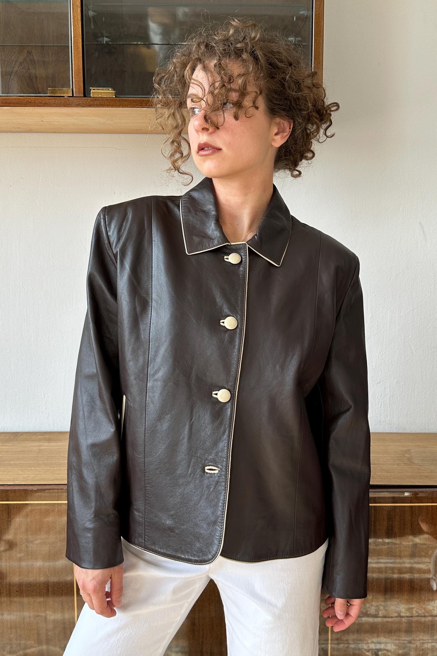 A woman with curly hair wearing a black leather jacket with white buttons poses in front of a glass display case.