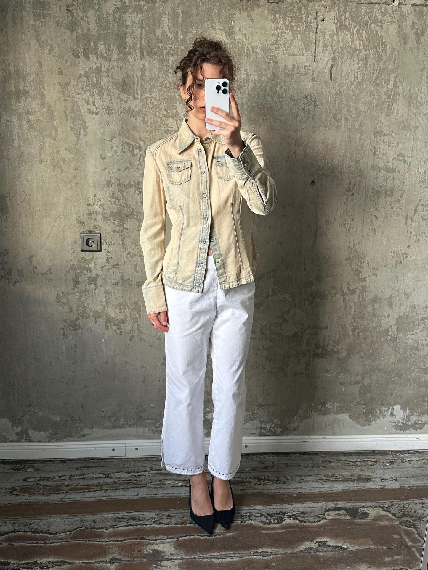 A woman is taking a selfie with a white phone, standing in front of a concrete wall.