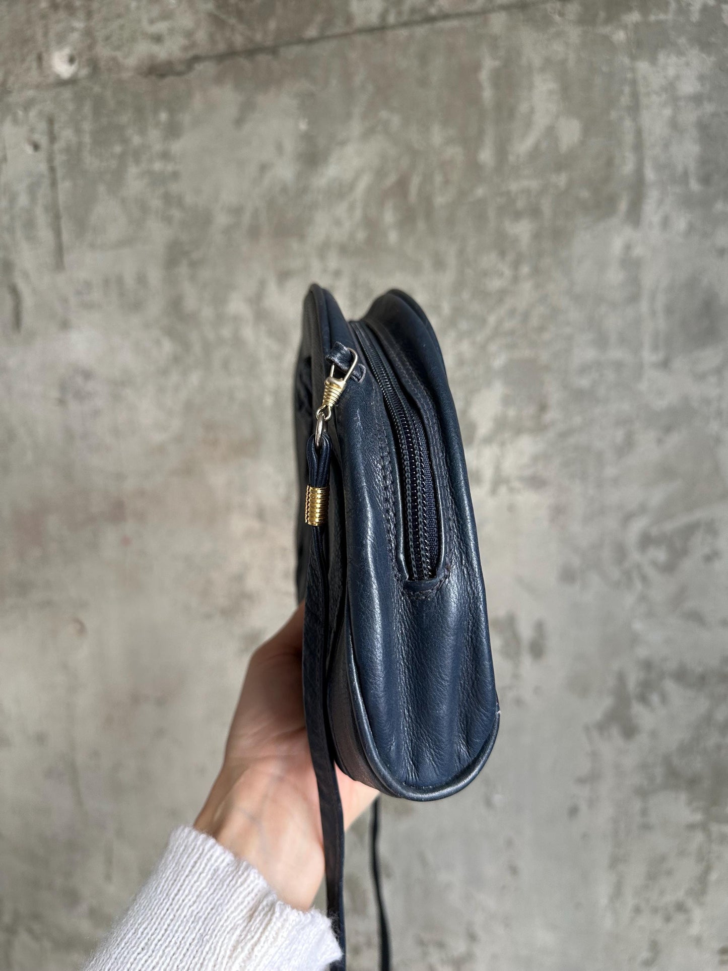 A hand holding a black leather crossbody bag with a zipper closure.