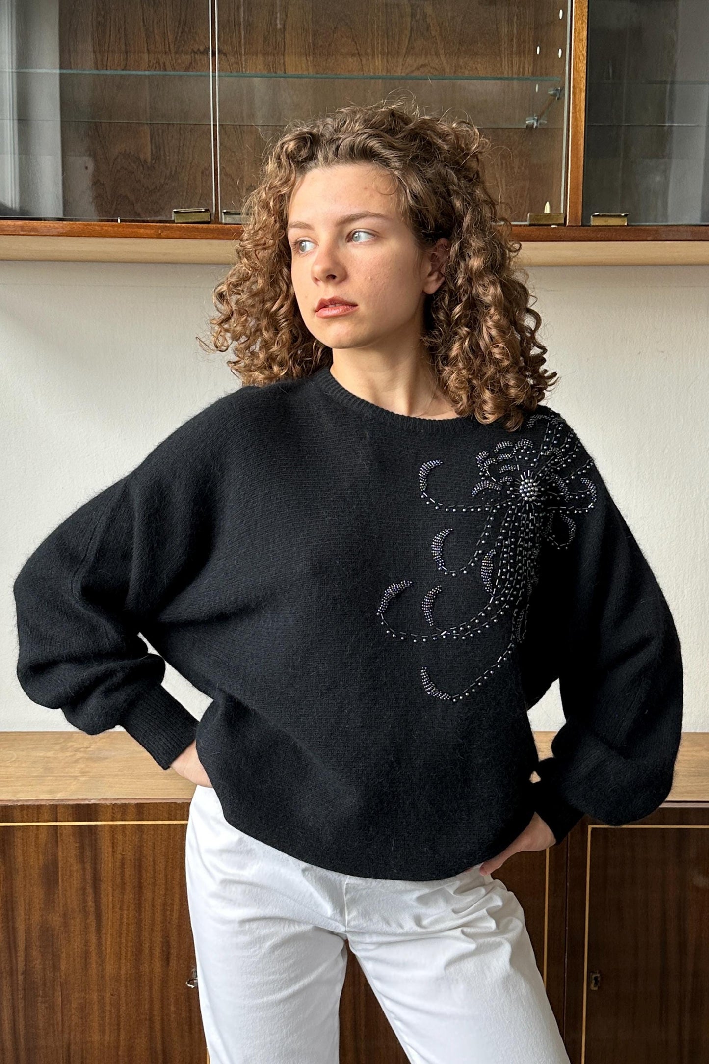 A woman with curly hair wearing a black sweater with a purple and white design stands in front of a wooden cabinet.