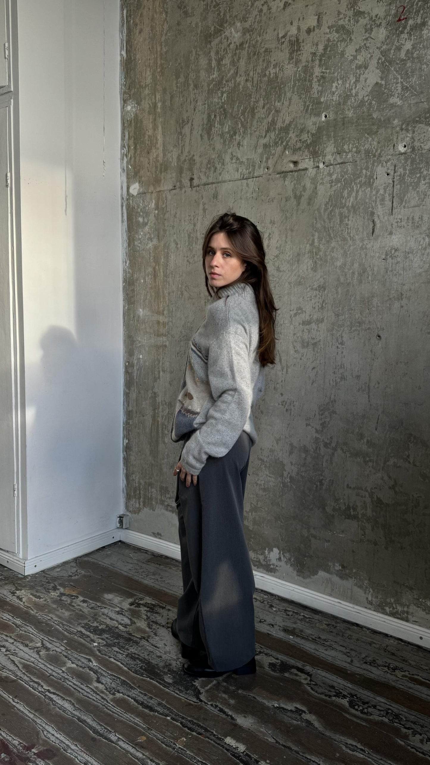 A woman in a gray sweater and dark pants stands in a room with a concrete wall and wooden floor.