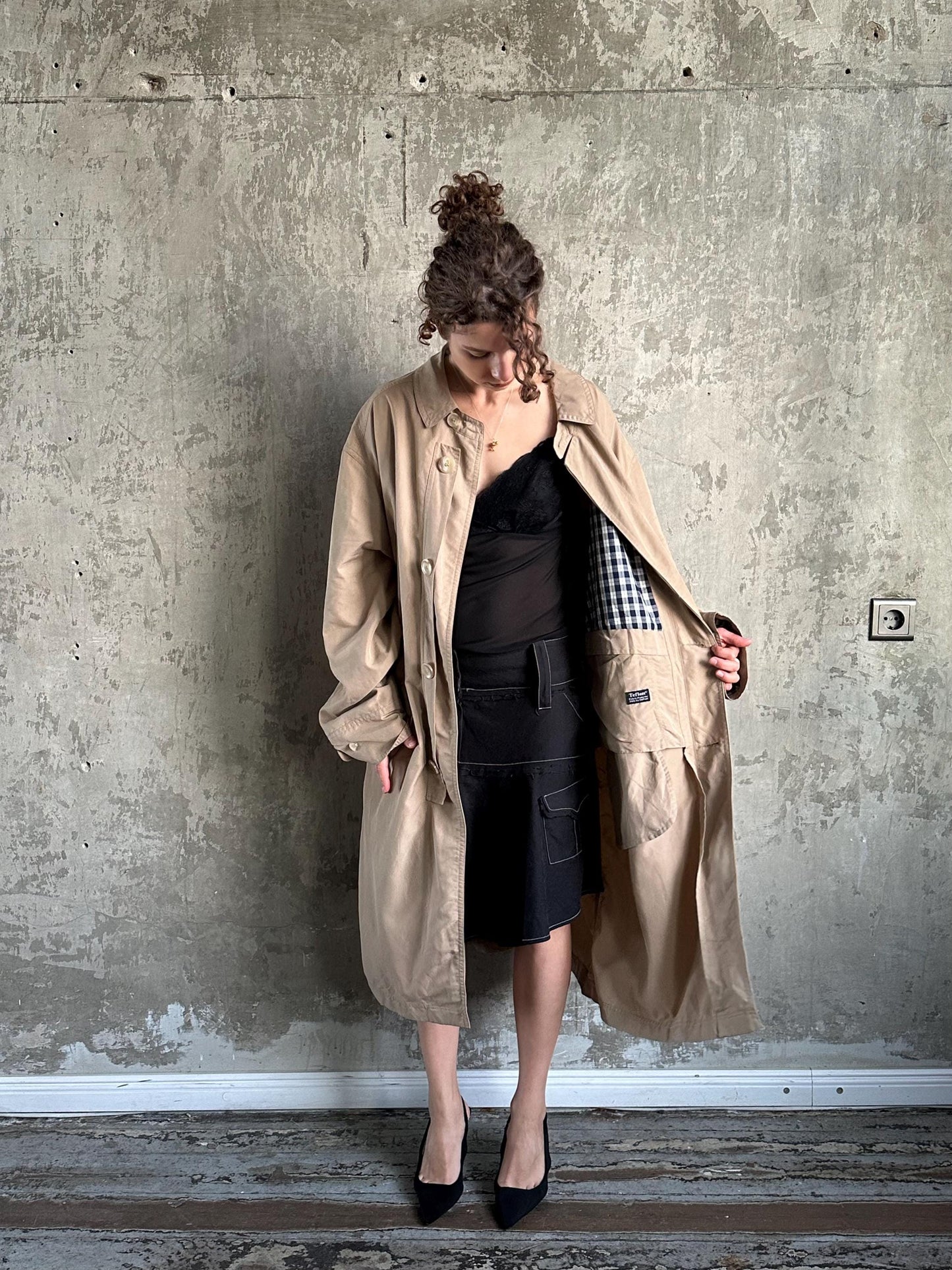 A woman in a beige trench coat stands against a concrete wall, her head turned to the side.