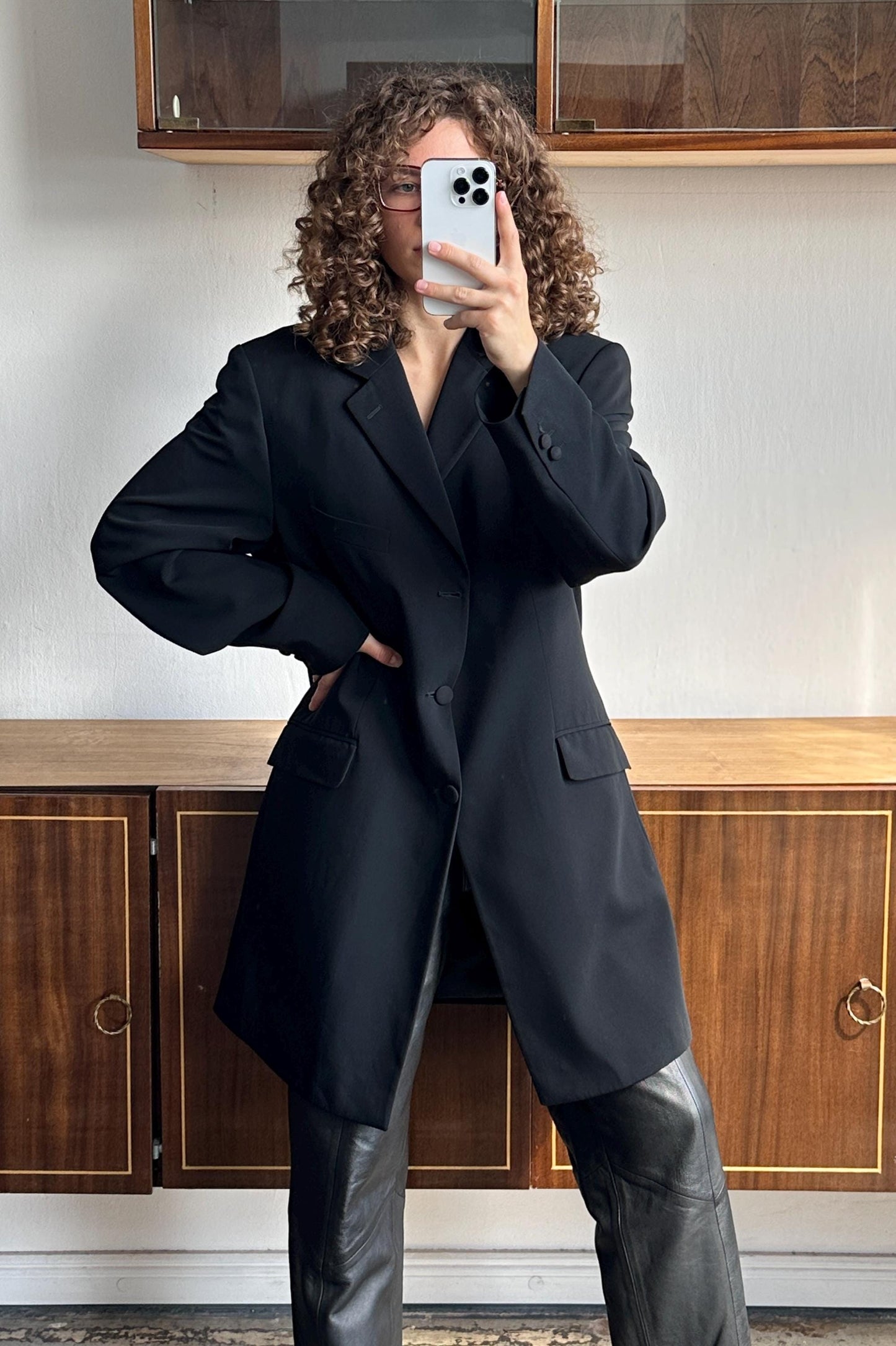 A woman with curly hair is taking a selfie with a white iPhone, wearing a black blazer and leather pants.