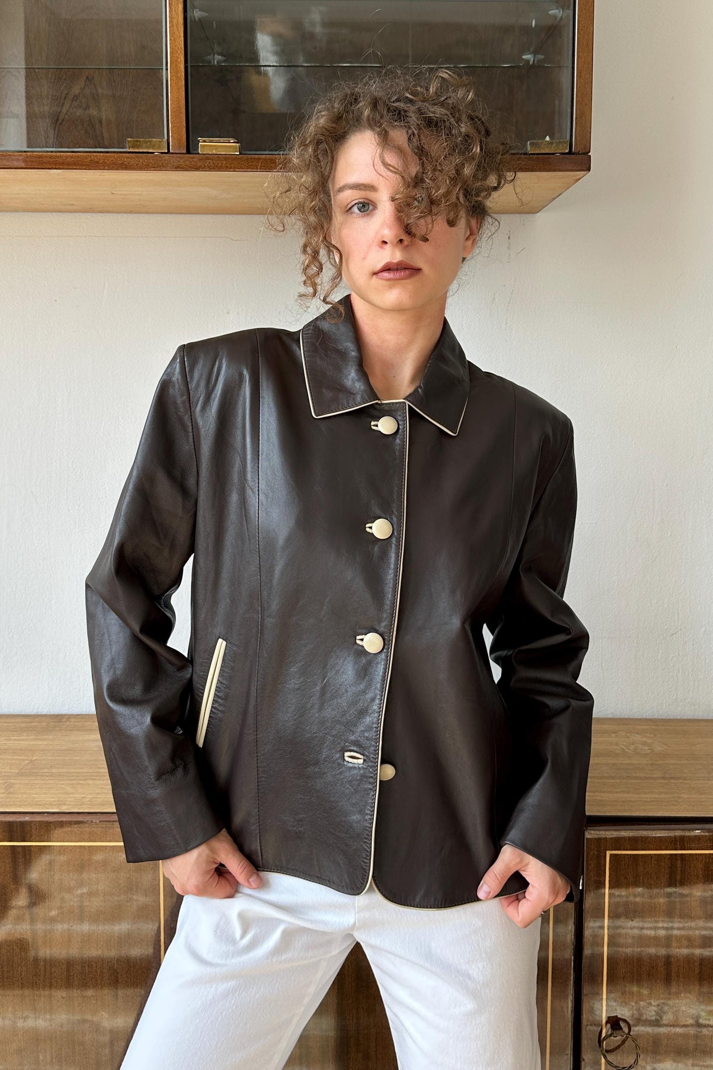 A woman wearing a black leather jacket with white buttons stands in front of a wooden cabinet.