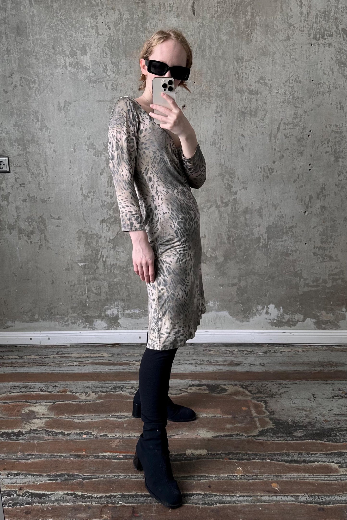 A person is taking a selfie with a smartphone, wearing a leopard print dress and sunglasses, standing in front of a concrete wall.