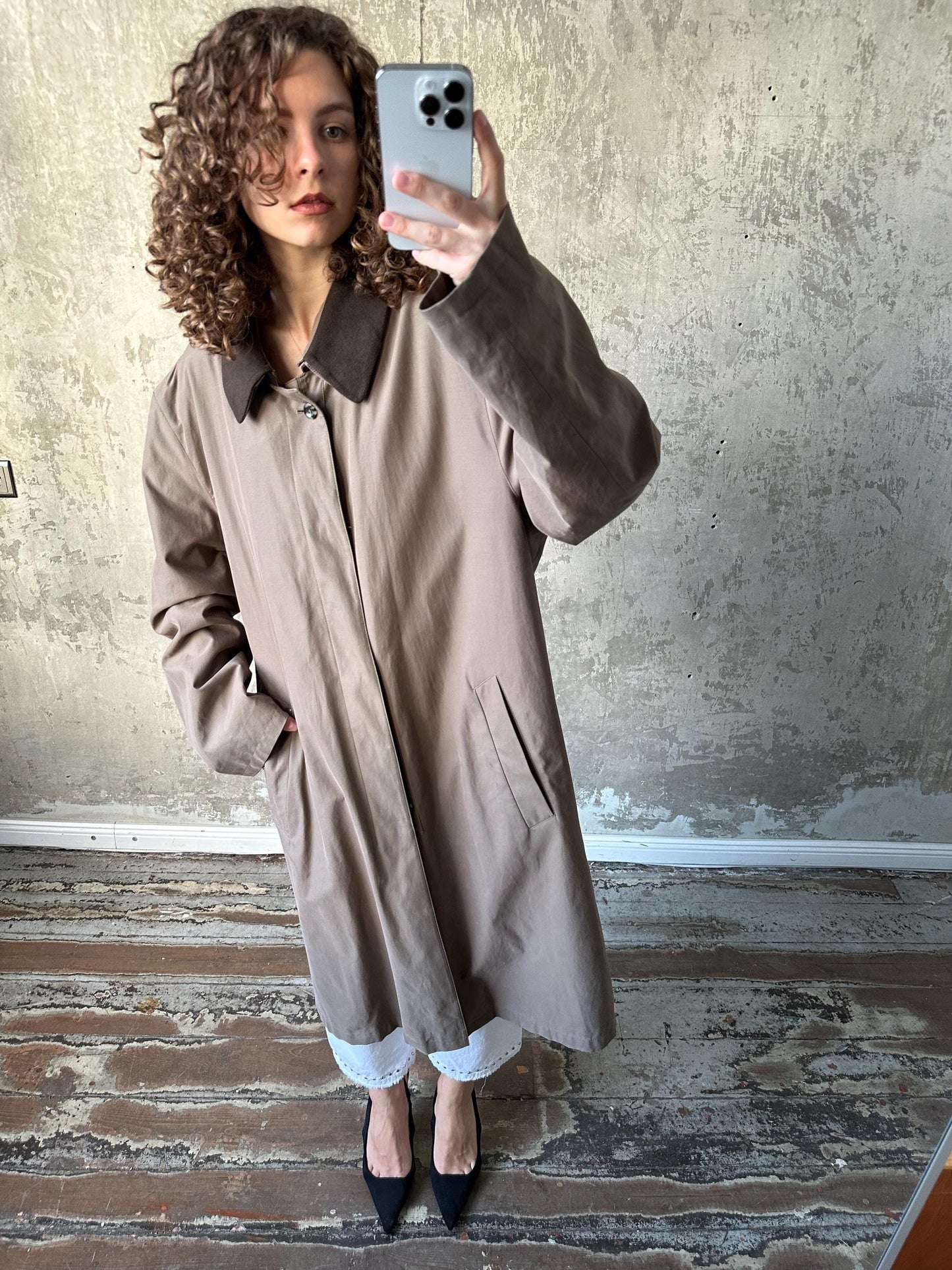 A woman with curly hair is taking a selfie with a smartphone, wearing a beige coat and standing in front of a concrete wall.