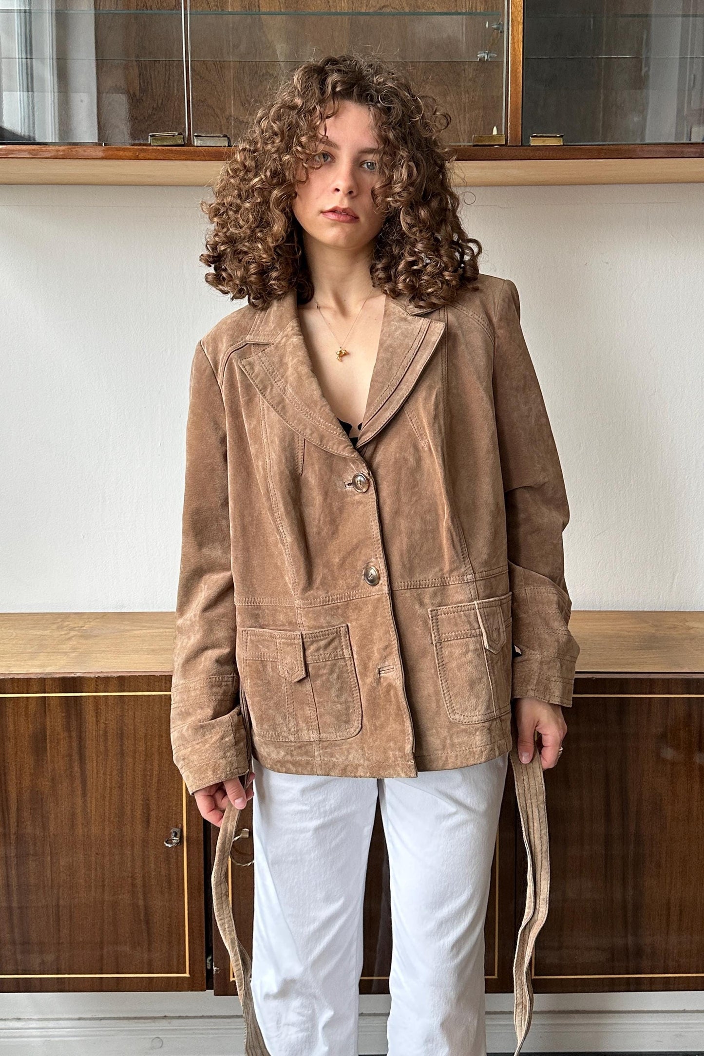 A woman with curly hair wearing a tan jacket and white pants stands in front of a wooden cabinet.