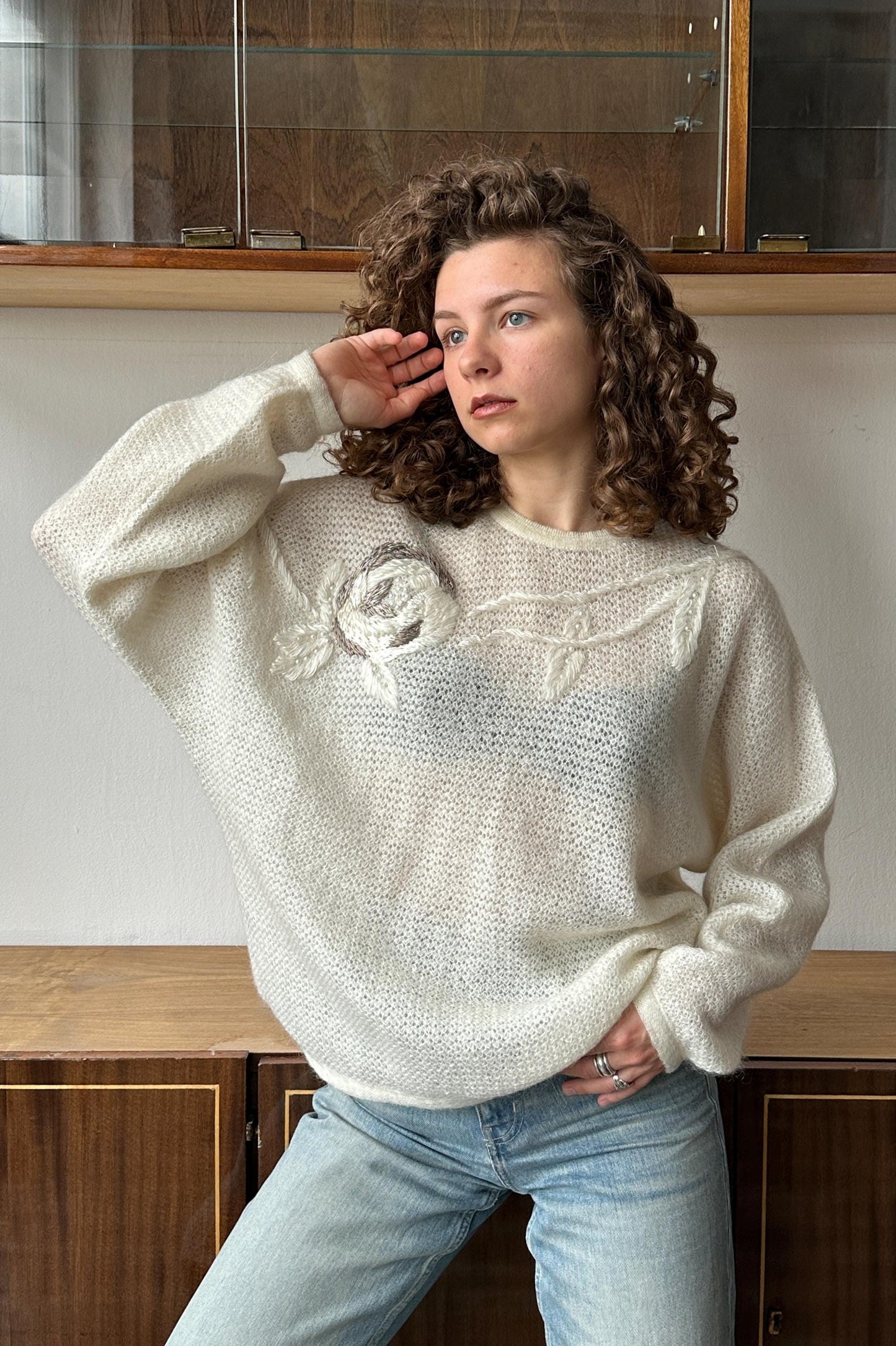 A young woman with curly hair wearing a white sweater with a floral design stands in front of a wooden cabinet.