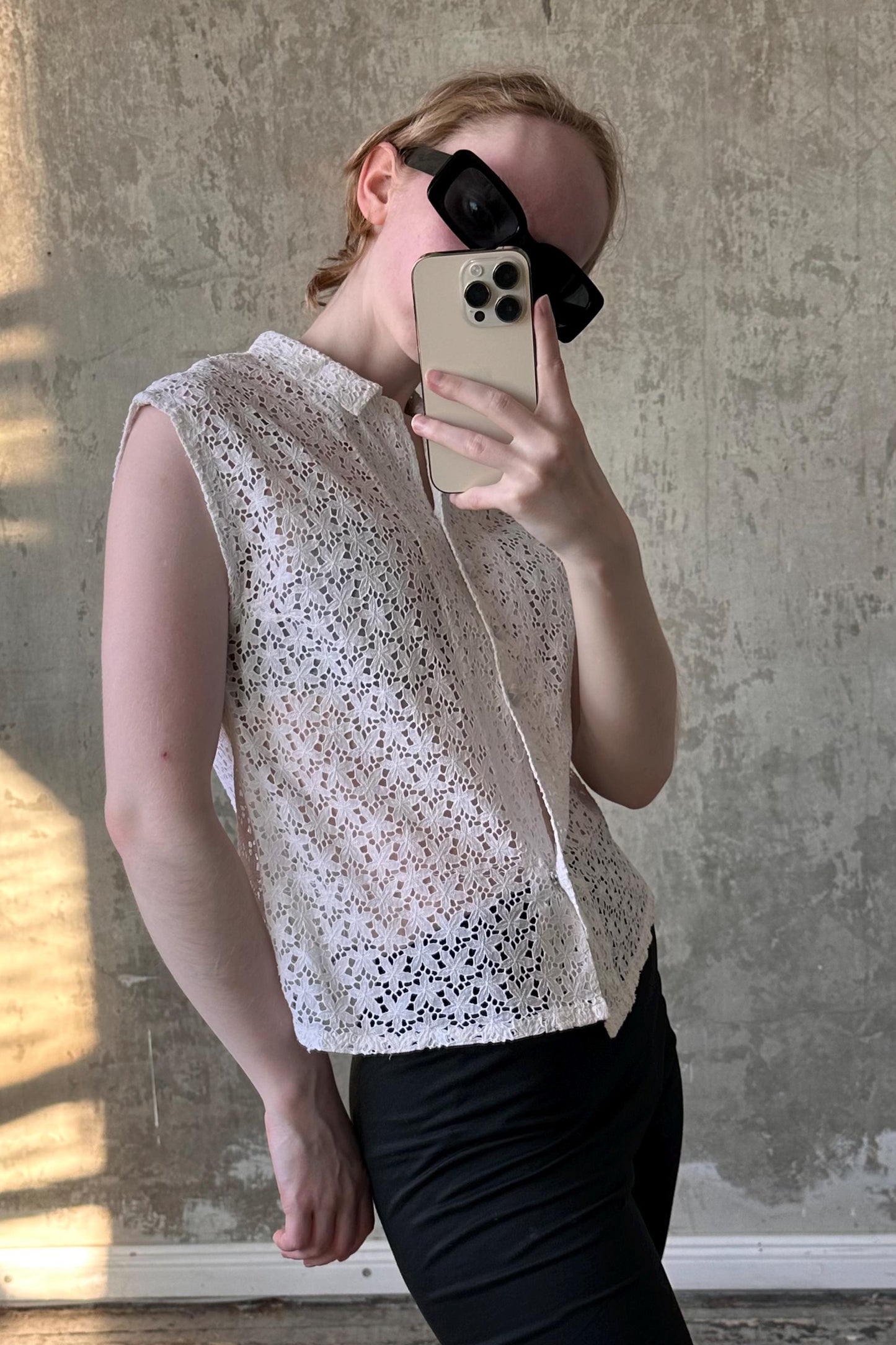 A person is taking a selfie with a smartphone, wearing a white lace top and black pants, and holding the phone up to their face.