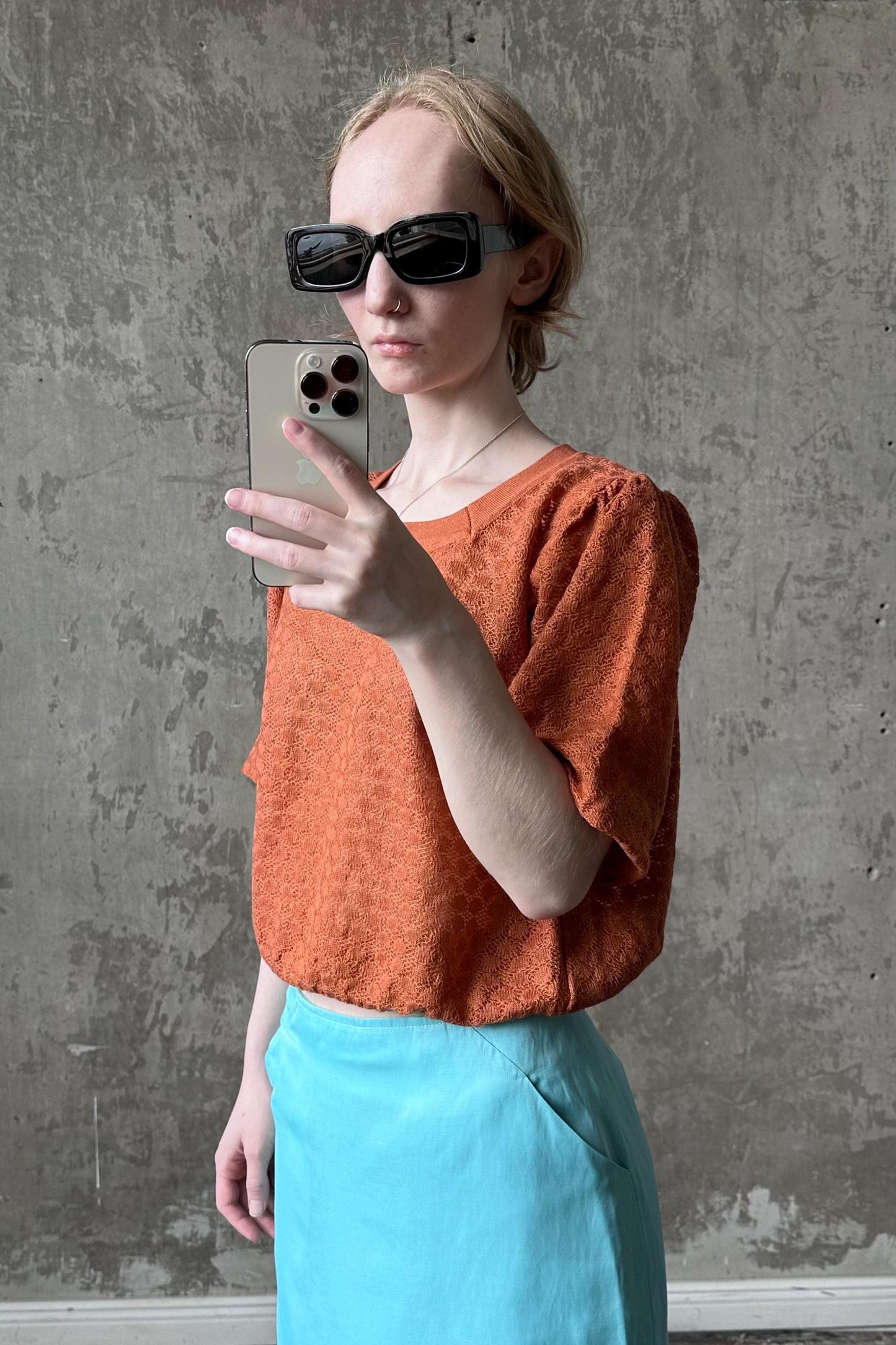 A woman wearing sunglasses and an orange top is taking a selfie with a smartphone.